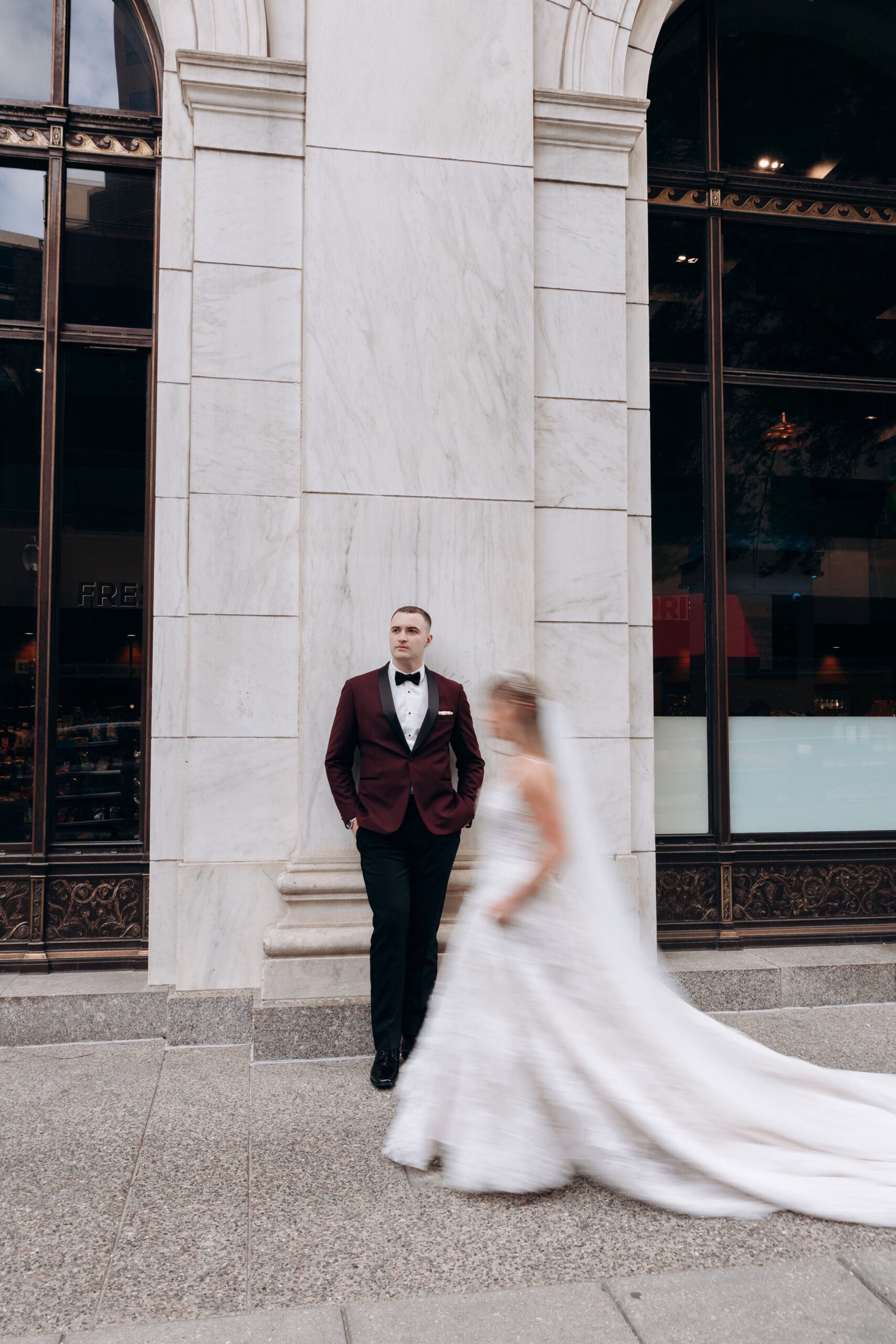Groom in burgundy tuxedo leaning against marble wall with blurred bride walking past in white wedding gown at Union Trust, Philadelphia.