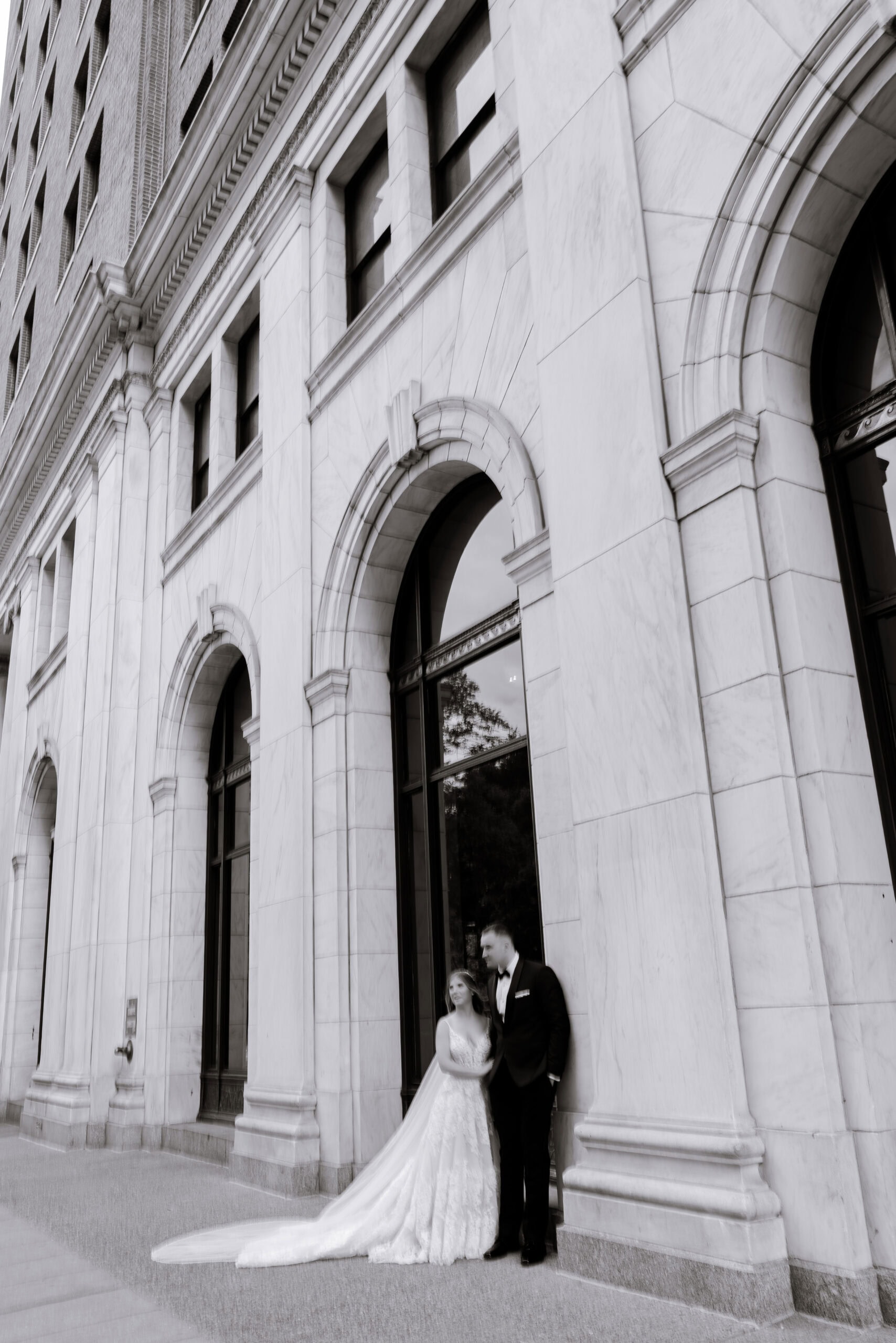 Bride in lace gown and groom in tuxedo standing beneath marble arches at Union Trust building, Philadelphia, PA.