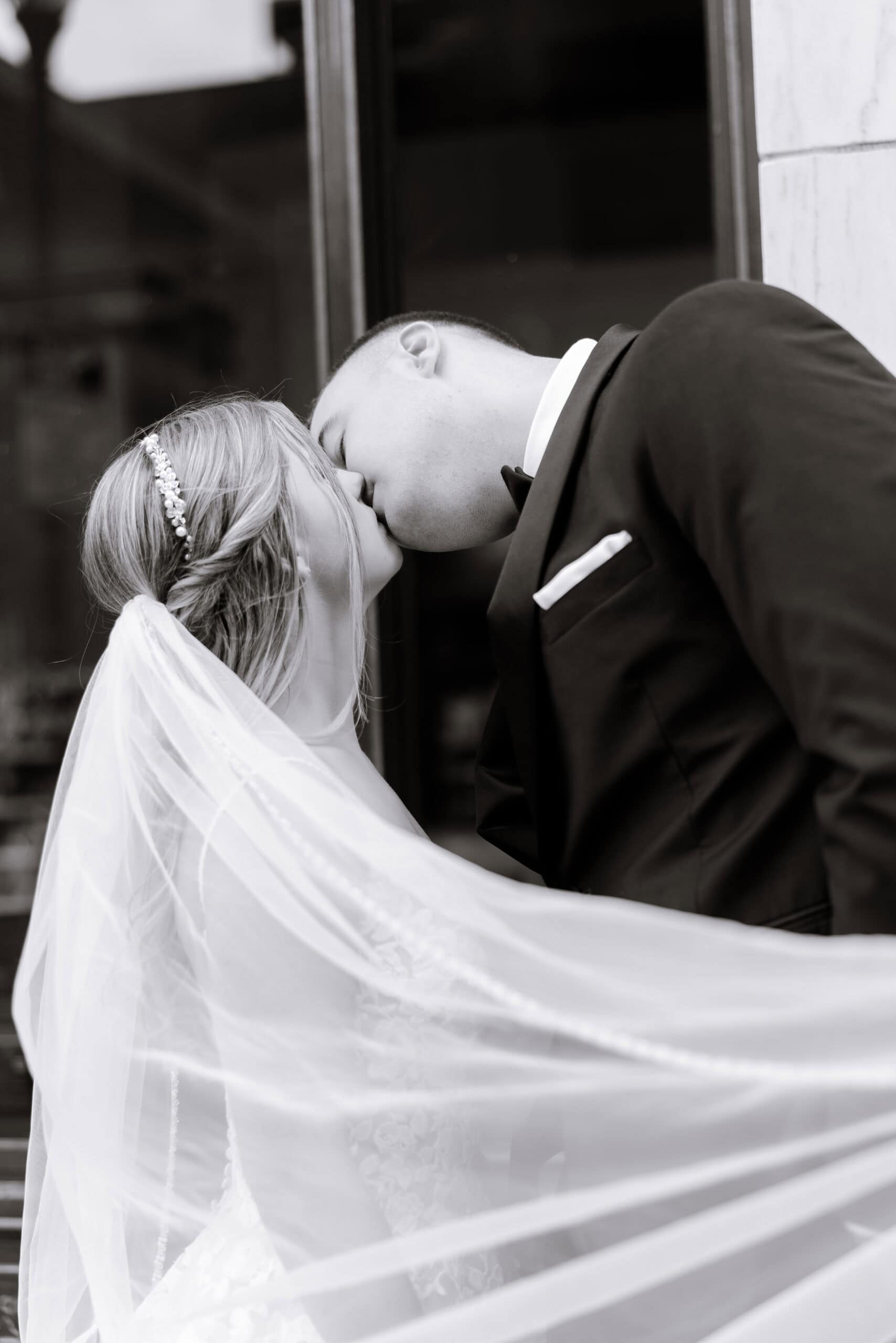 Black and white photo of a bride and groom sharing a kiss, the bride's veil flowing and the groom in a tuxedo, Union Trust venue.