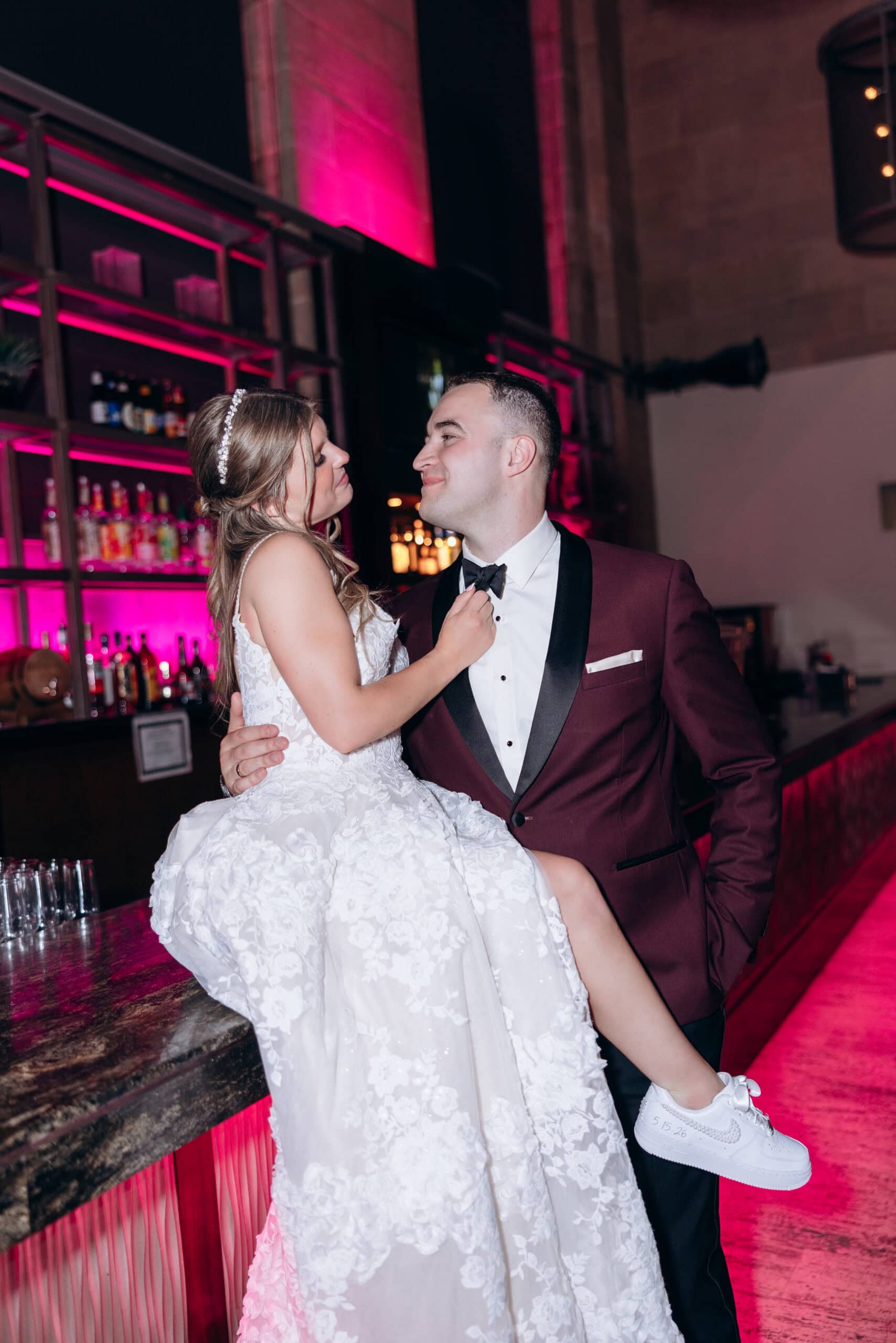 Bride in floral lace wedding dress and custom white sneakers being held by groom in burgundy tuxedo near a pink-lit bar at Union Trust, Philadelphia.