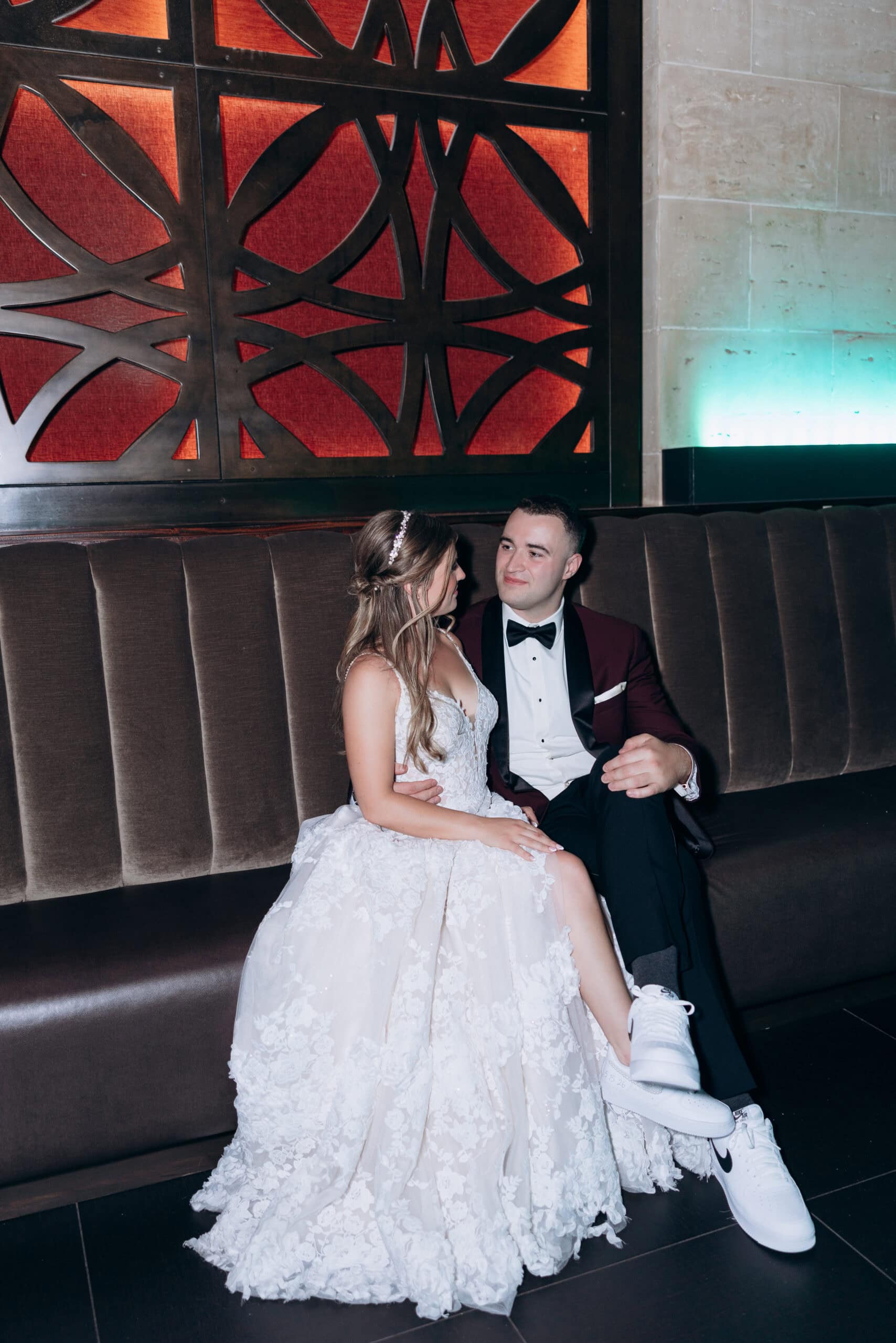 Bride in lace gown and groom in tuxedo with white sneakers seated on velvet banquette under intricate metal wall panel at Union Trust, Philadelphia.