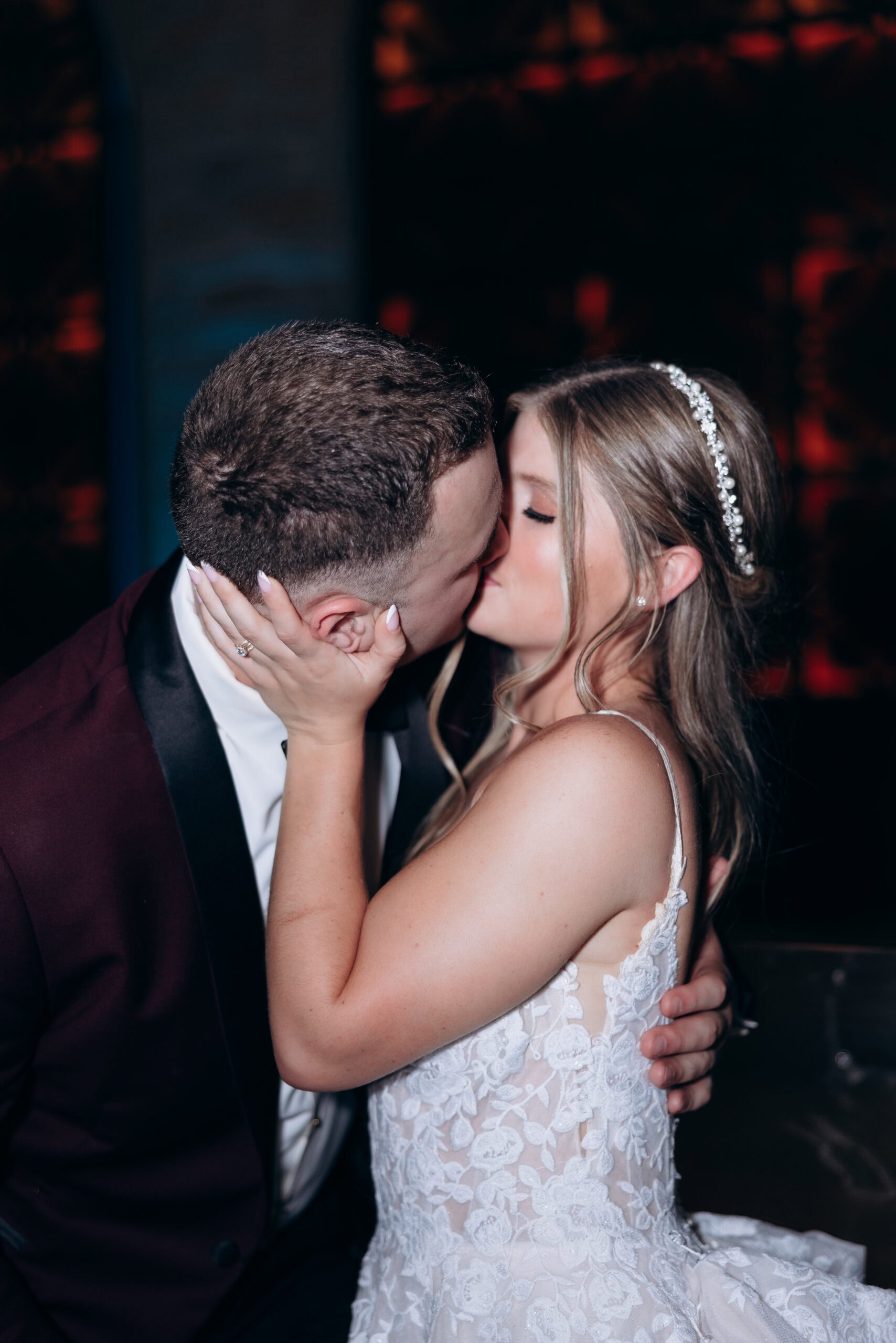 Bride and groom sharing a close kiss, bride wearing lace wedding dress and pearl headband, groom in dark tuxedo jacket, dark background.
