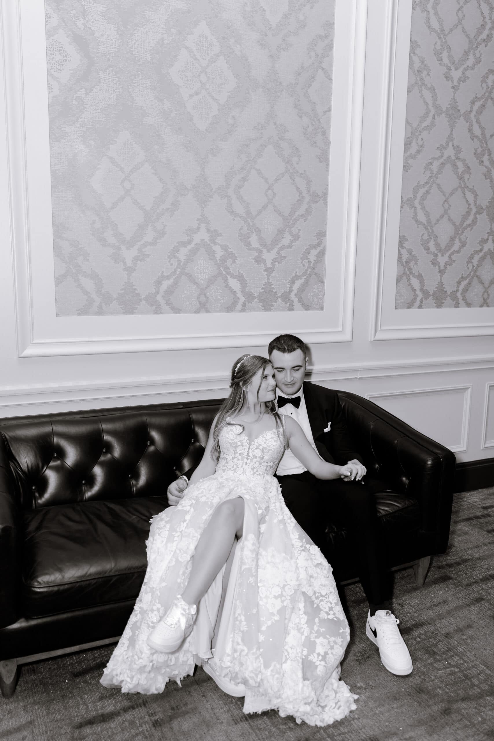 Bride in lace wedding dress and sneakers leaning on groom in tuxedo sitting on black leather sofa with patterned wall panels behind them at Union Trust, Philadelphia.