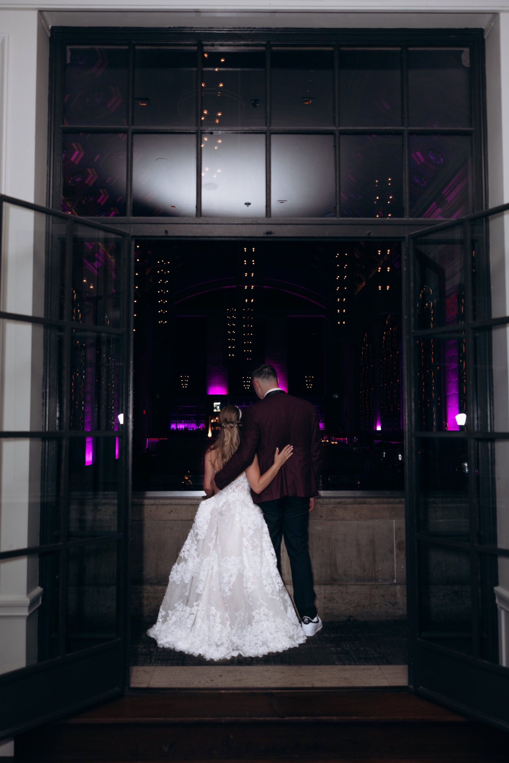 Bride in floral lace gown and groom in dark suit stand arm in arm, viewed from behind through open glass doors at Union Trust venue with purple lighting.