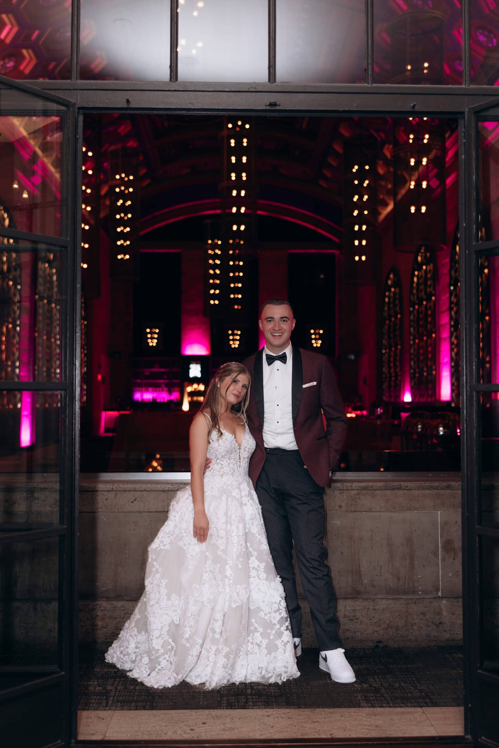 Bride in floral lace wedding dress and groom in burgundy tuxedo jacket standing in an open window frame with pink-lit chandeliers and arched windows in the background at Union Trust, Philadelphia.