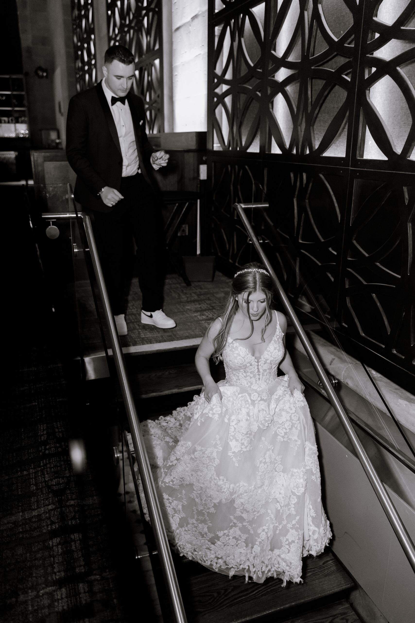 Bride in detailed floral lace wedding dress descending staircase, groom in tuxedo and white sneakers following behind, intricate geometric wall panels in background at Union Trust, Philadelphia.