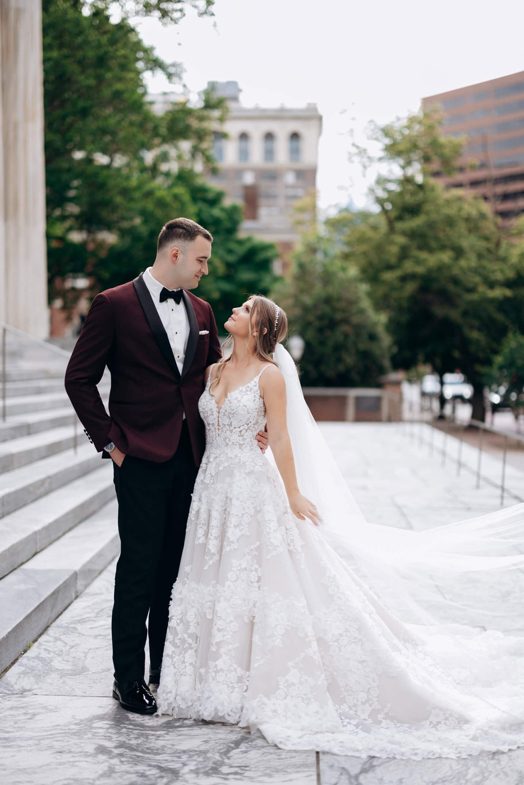 Bride in floral lace wedding gown and groom in burgundy tuxedo looking at each other on marble steps outside Union Trust, Philadelphia.