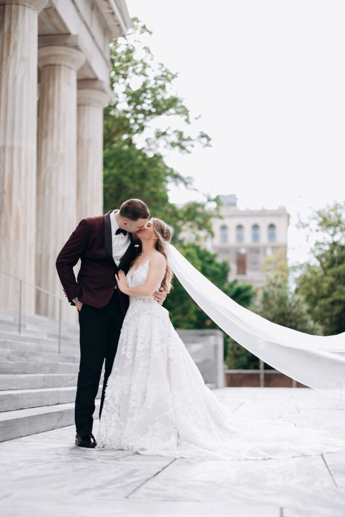 Bride and groom sharing a kiss on marble steps with classical columns and greenery in the background at Union Trust, Philadelphia.