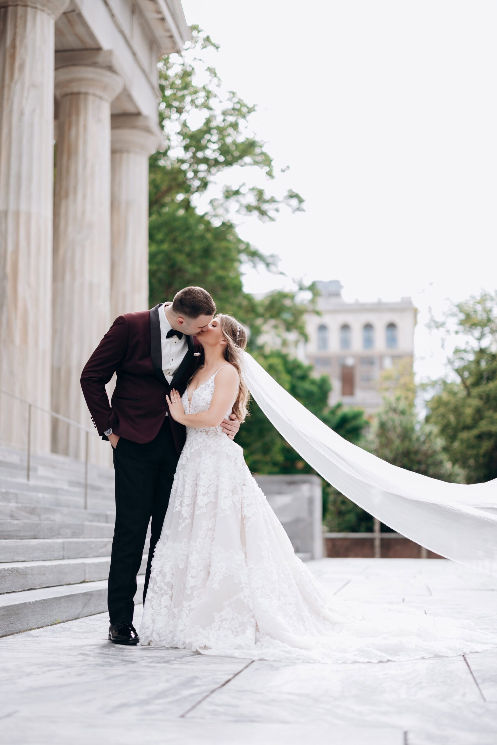 Bride and groom sharing a kiss on marble steps with classical columns and greenery in the background at Union Trust, Philadelphia.