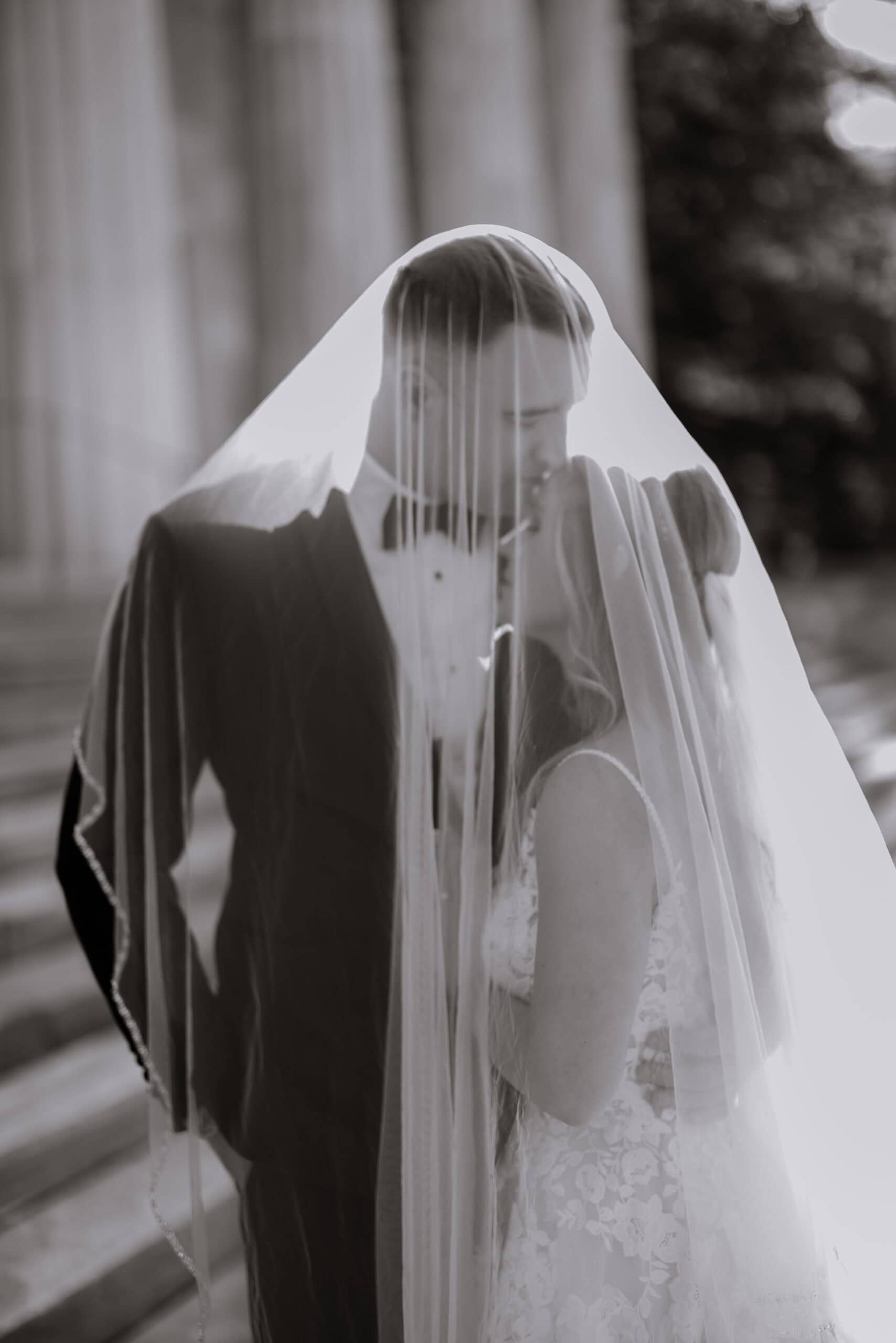Black and white photo of a bride and groom sharing an intimate moment under a veil, with the groom in a tuxedo and bride in lace gown at Union Trust, Philadelphia.