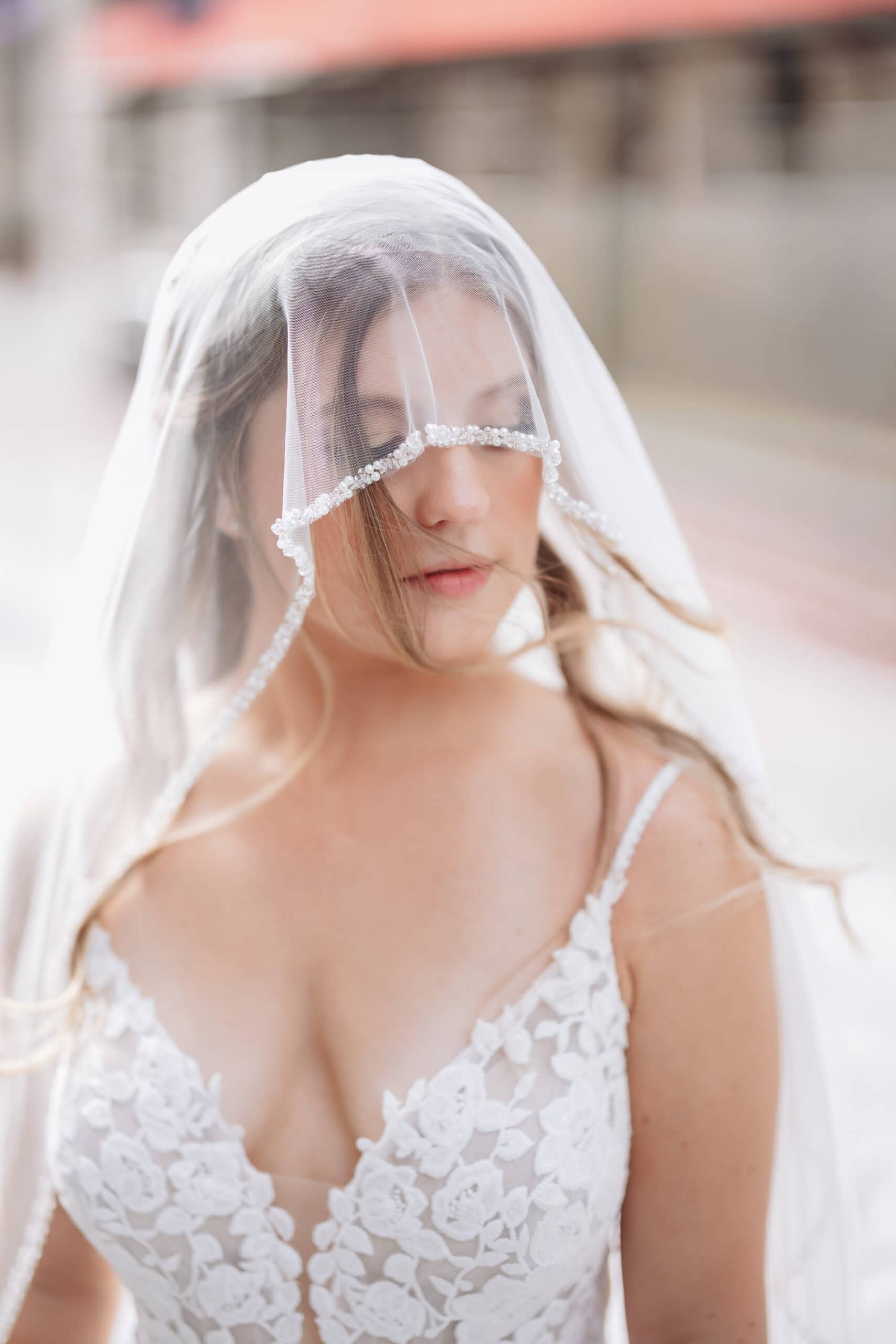 Close-up of a bride with eyes closed under a sheer veil with pearl trim, wearing a floral lace wedding dress.
