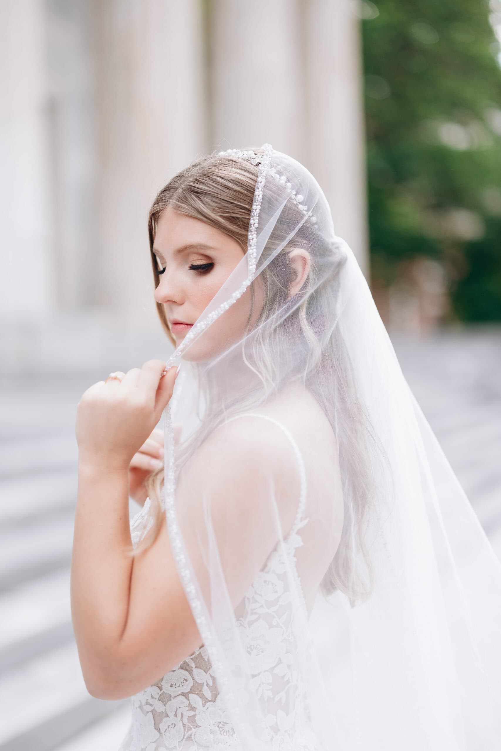 Close-up of a bride gently holding a beaded veil over her lace wedding dress, standing outside Union Trust in Philadelphia.