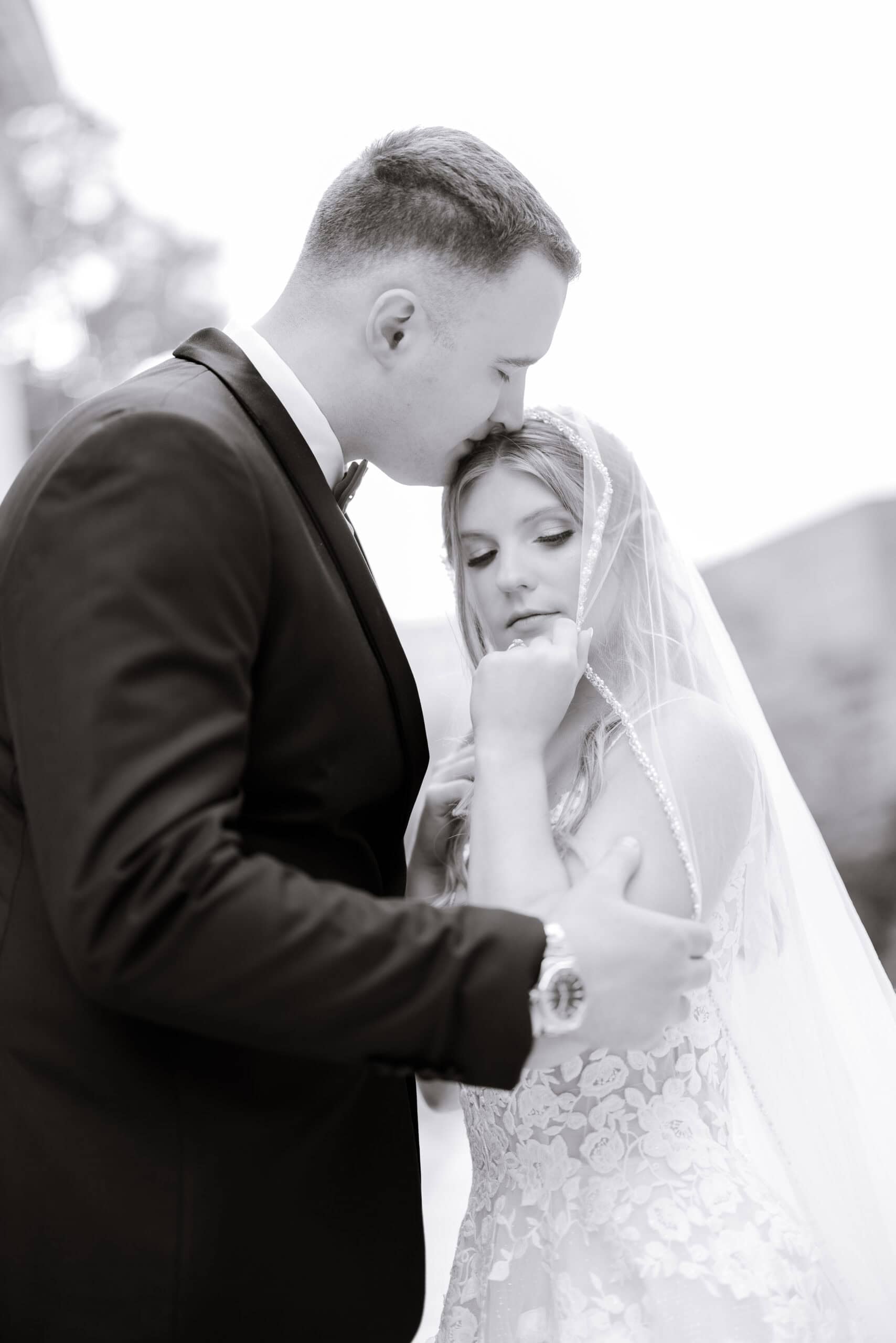 Black and white close-up of groom in tuxedo kissing bride's forehead while she holds veil, showcasing lace gown details at Union Trust, Philadelphia.