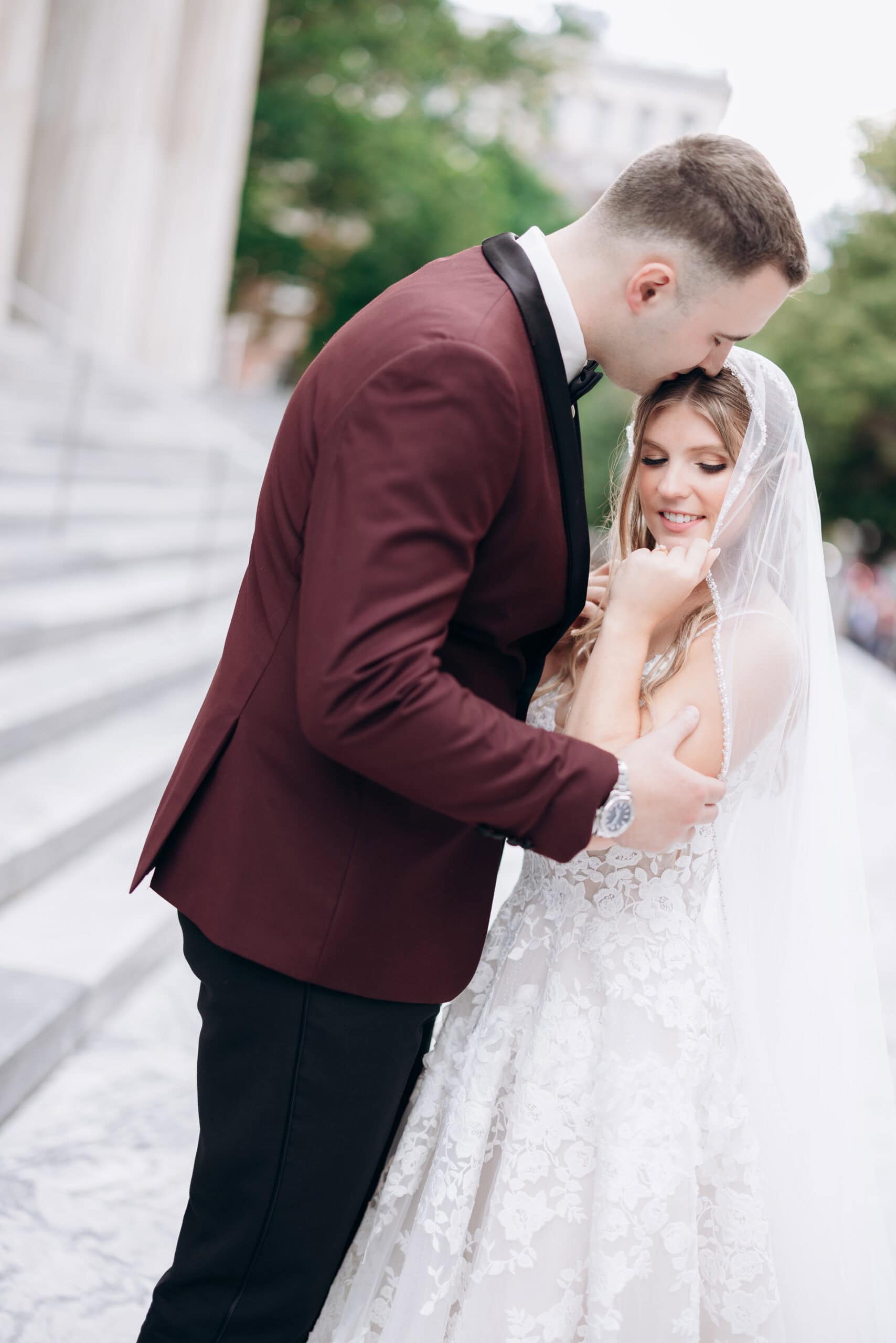 Groom in burgundy tuxedo kissing bride's forehead while she holds lace veil, steps and greenery blurred in background at Union Trust Philadelphia.