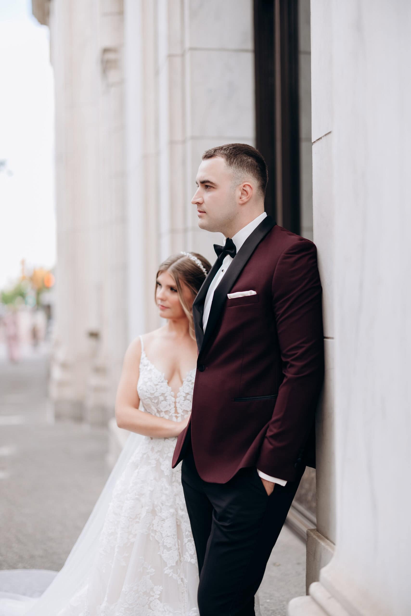 Groom in burgundy tuxedo leaning against marble column with bride in lace wedding dress behind him at Union Trust, Philadelphia.