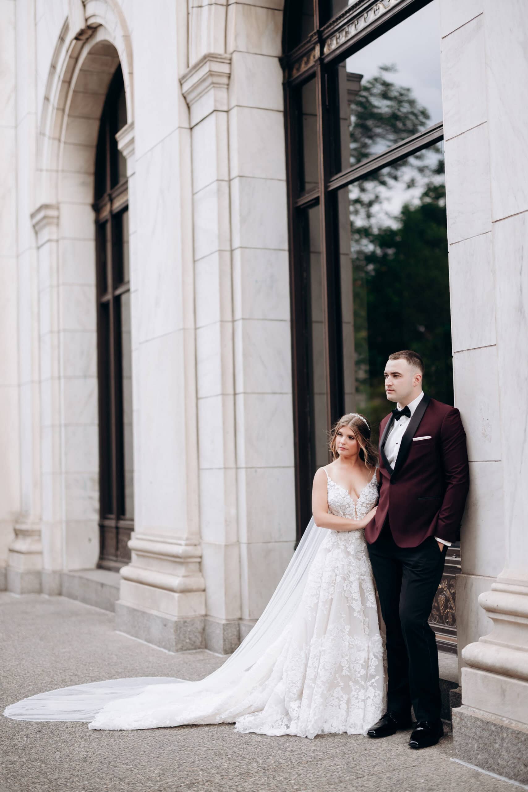 Bride in lace wedding dress with long train leaning on groom in burgundy tuxedo outside marble building at Union Trust Philadelphia.