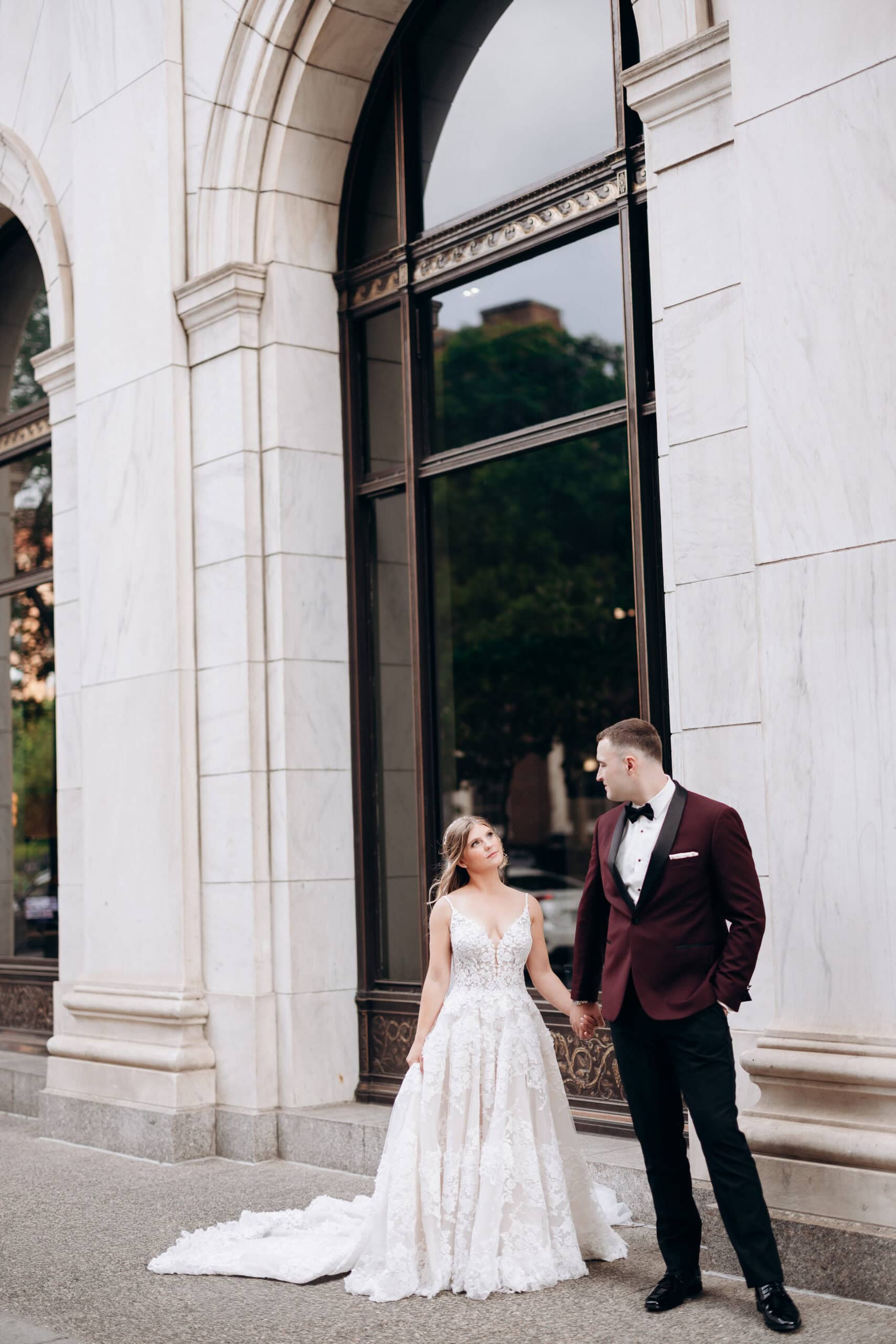 Bride in lace wedding dress and groom in burgundy tuxedo holding hands under marble arches at Union Trust, Philadelphia.