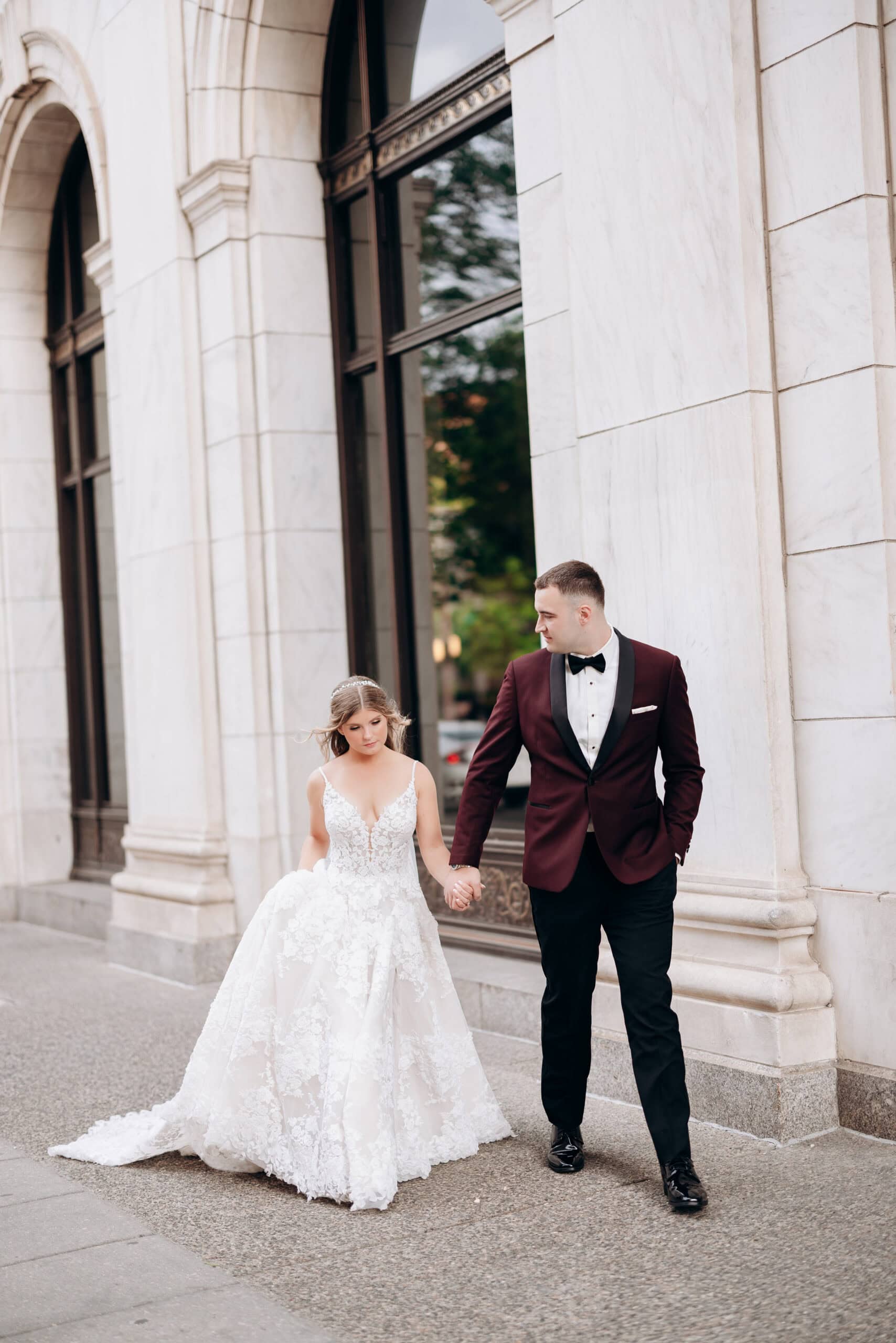 Bride in lace wedding gown and groom in burgundy tuxedo holding hands walking outside marble building at Union Trust, Philadelphia.