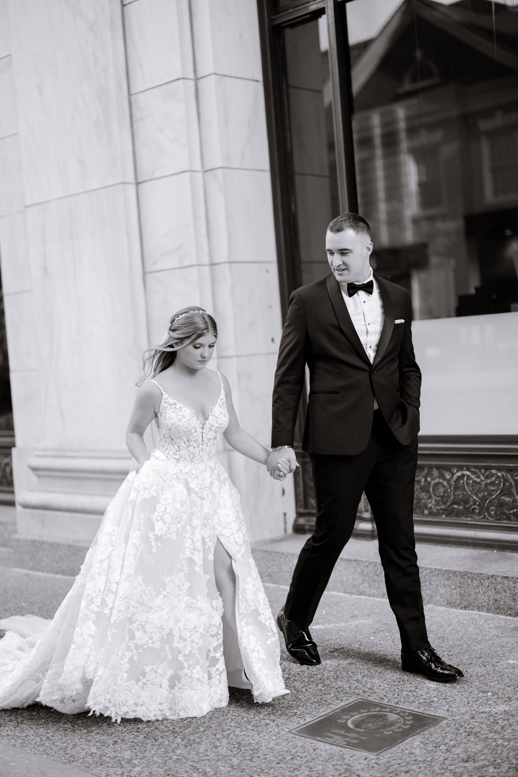 Bride in floral lace wedding dress and groom in black tuxedo holding hands, walking past marble columns at Union Trust, Philadelphia.