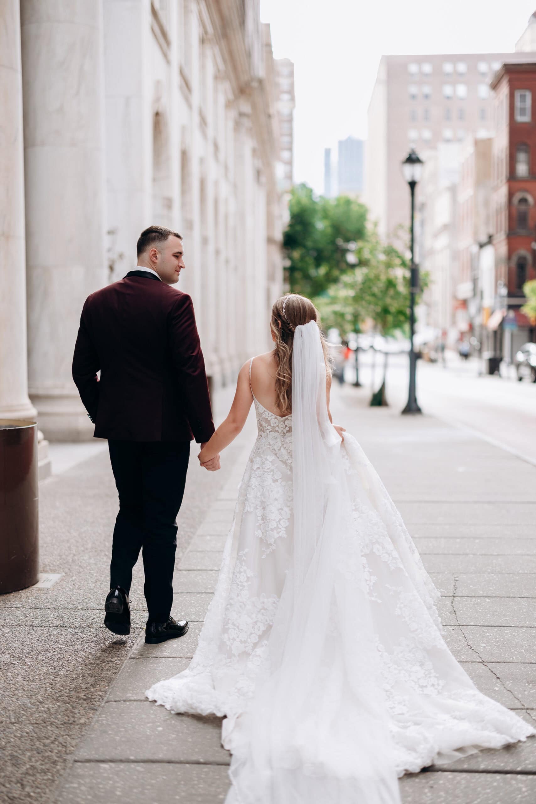 Bride and groom walking hand in hand on a city sidewalk with marble columns and urban buildings in the background, Philadelphia Union Trust venue.