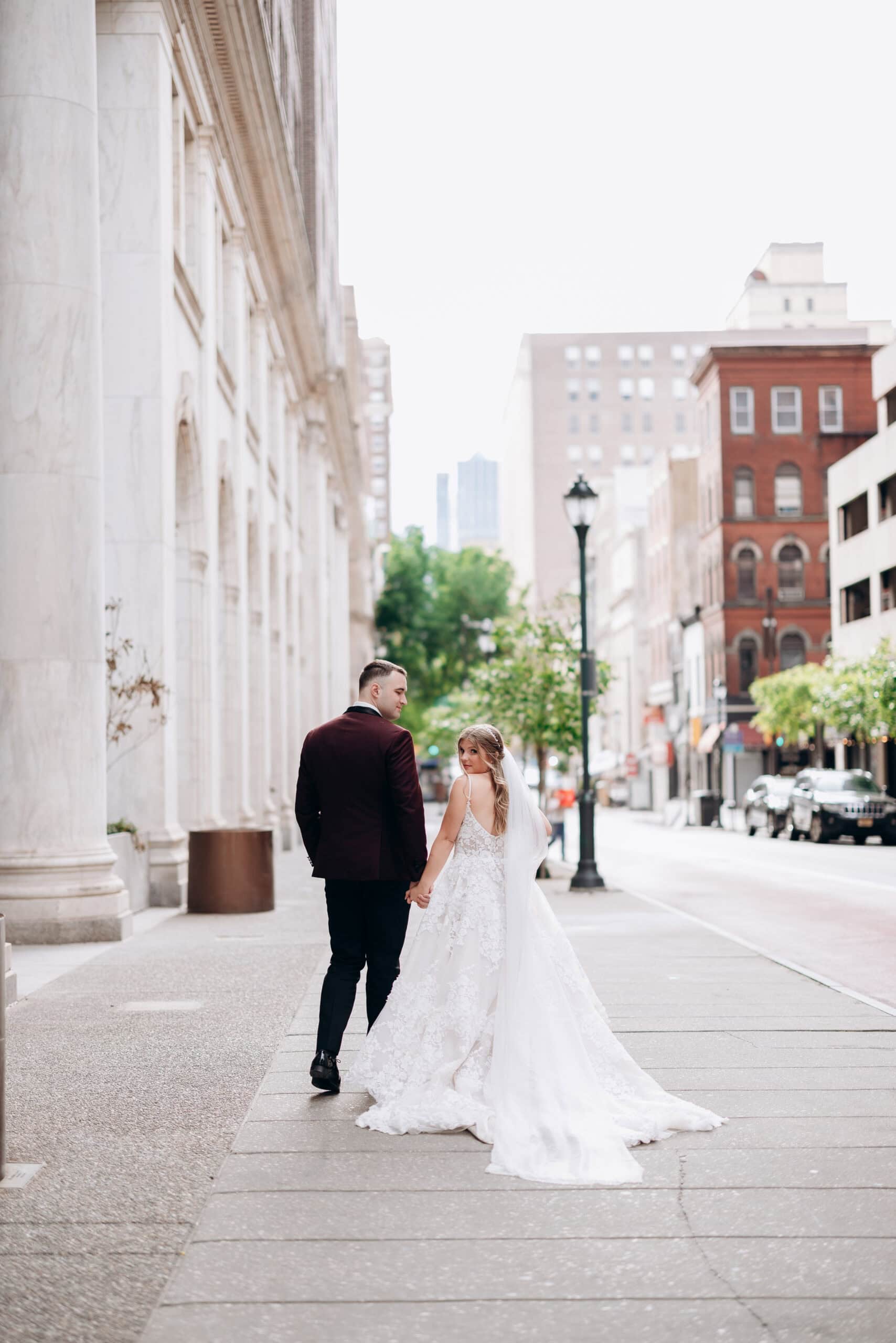 Bride and groom holding hands walking down a city sidewalk with marble columns and historic buildings in Philadelphia, PA.