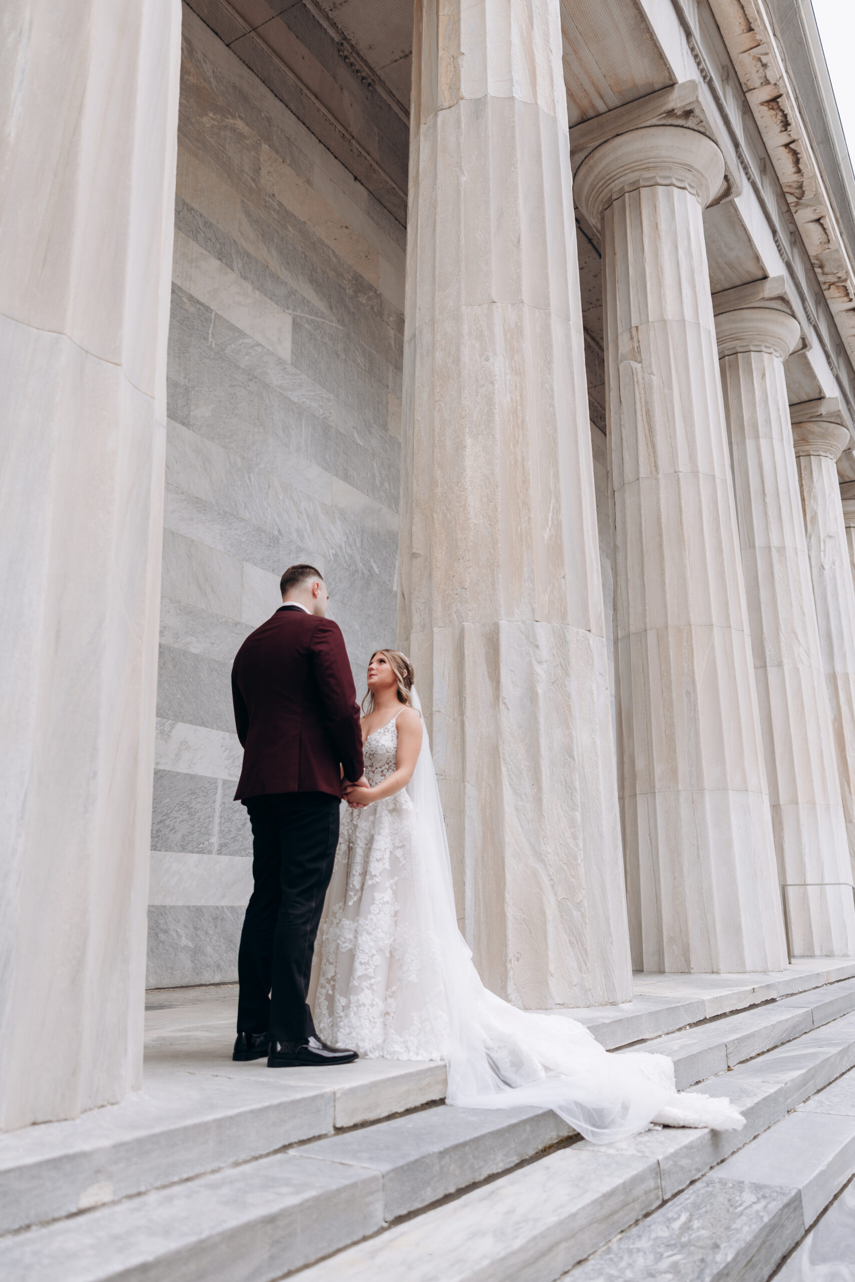 Bride and groom holding hands on marble steps beneath large classical columns at Union Trust, Philadelphia.