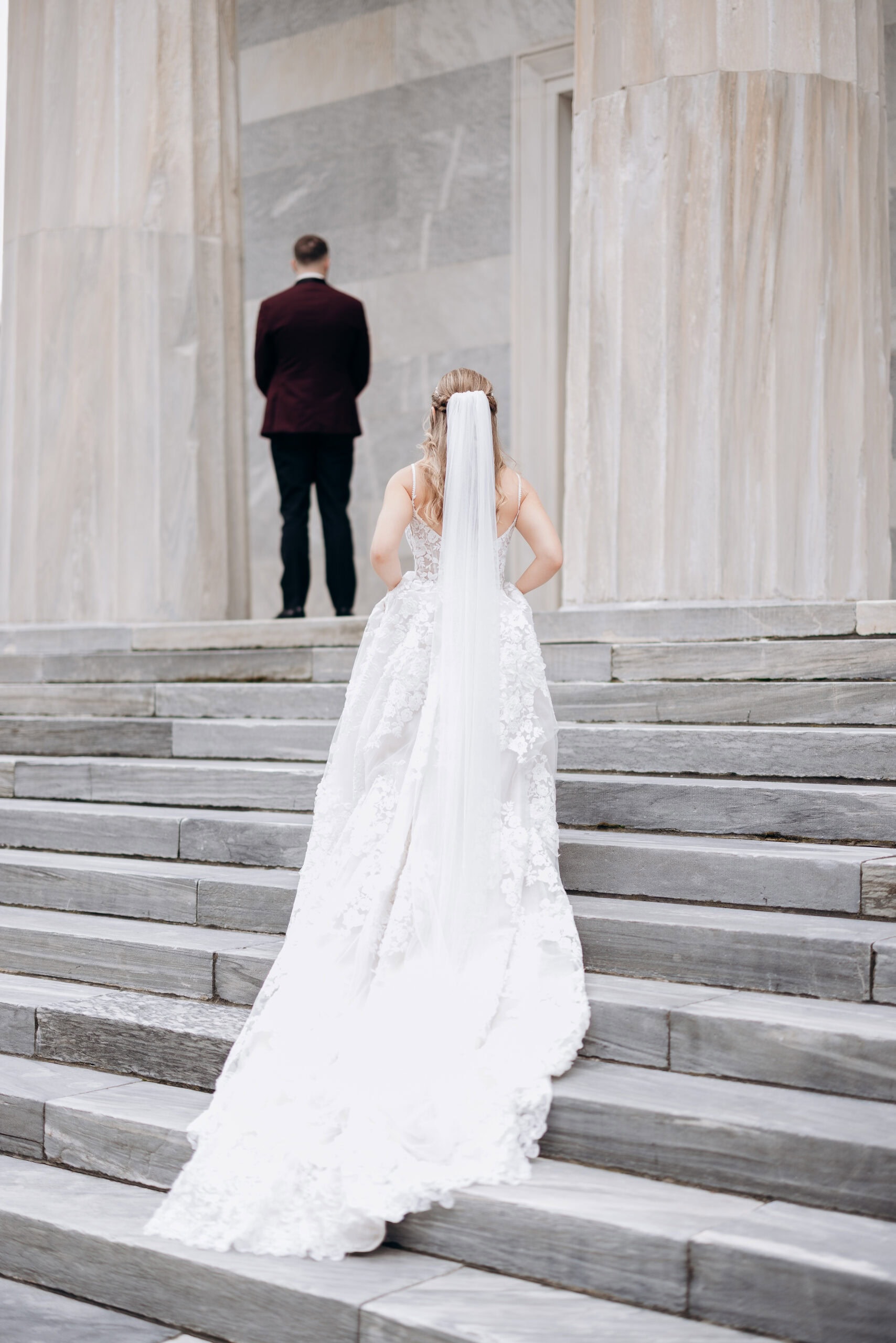Bride in detailed lace wedding dress with long veil ascending marble steps toward groom in burgundy jacket standing with back turned at Union Trust, Philadelphia.