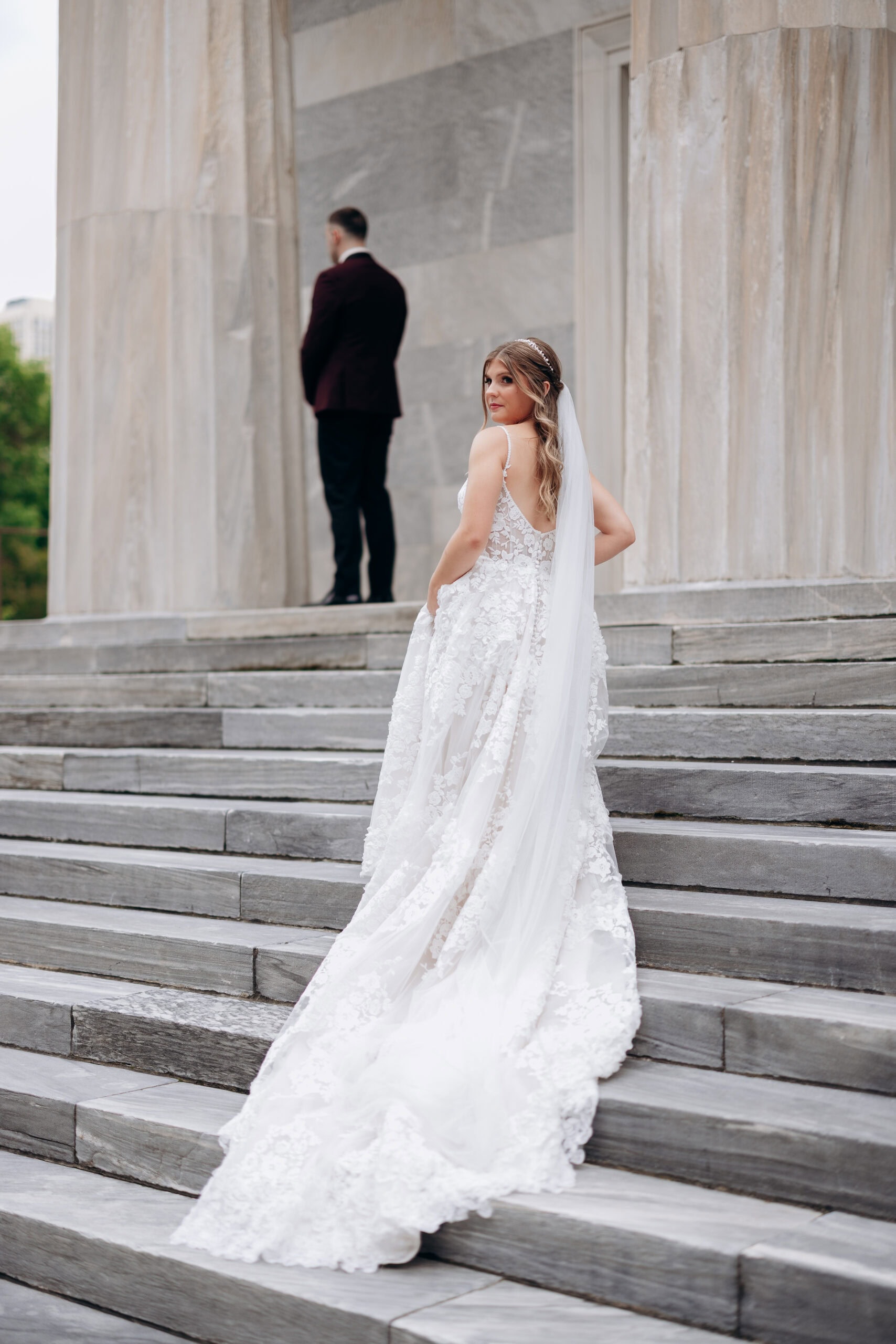Bride in lace wedding dress with long veil on marble steps, groom in dark suit facing away, Union Trust Philadelphia columns in background.