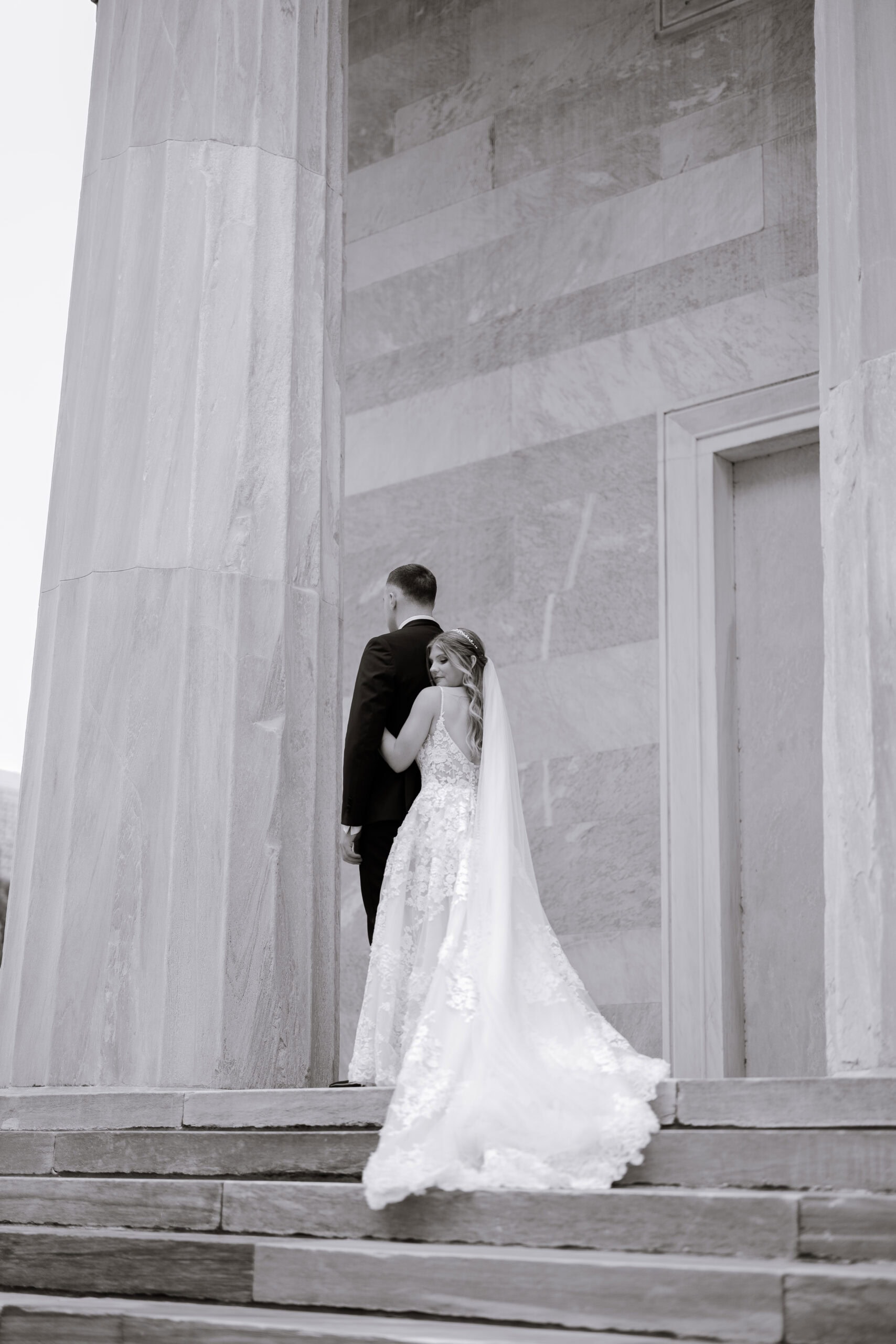Bride in lace wedding dress and veil hugging groom in black suit on stone steps by large marble columns at Union Trust, Philadelphia.