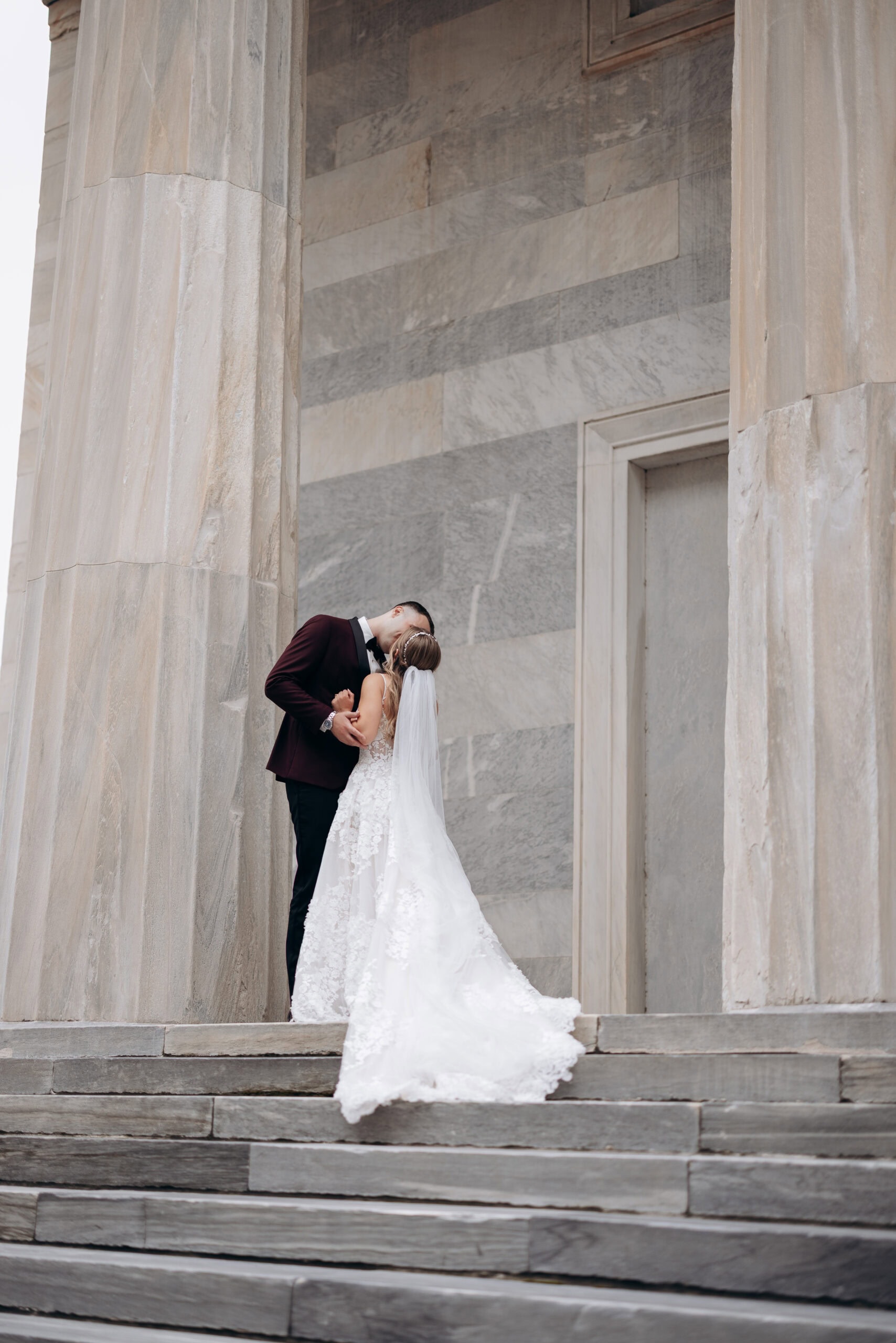 Bride and groom sharing a kiss on stone steps between large marble columns at Union Trust, Philadelphia, PA.