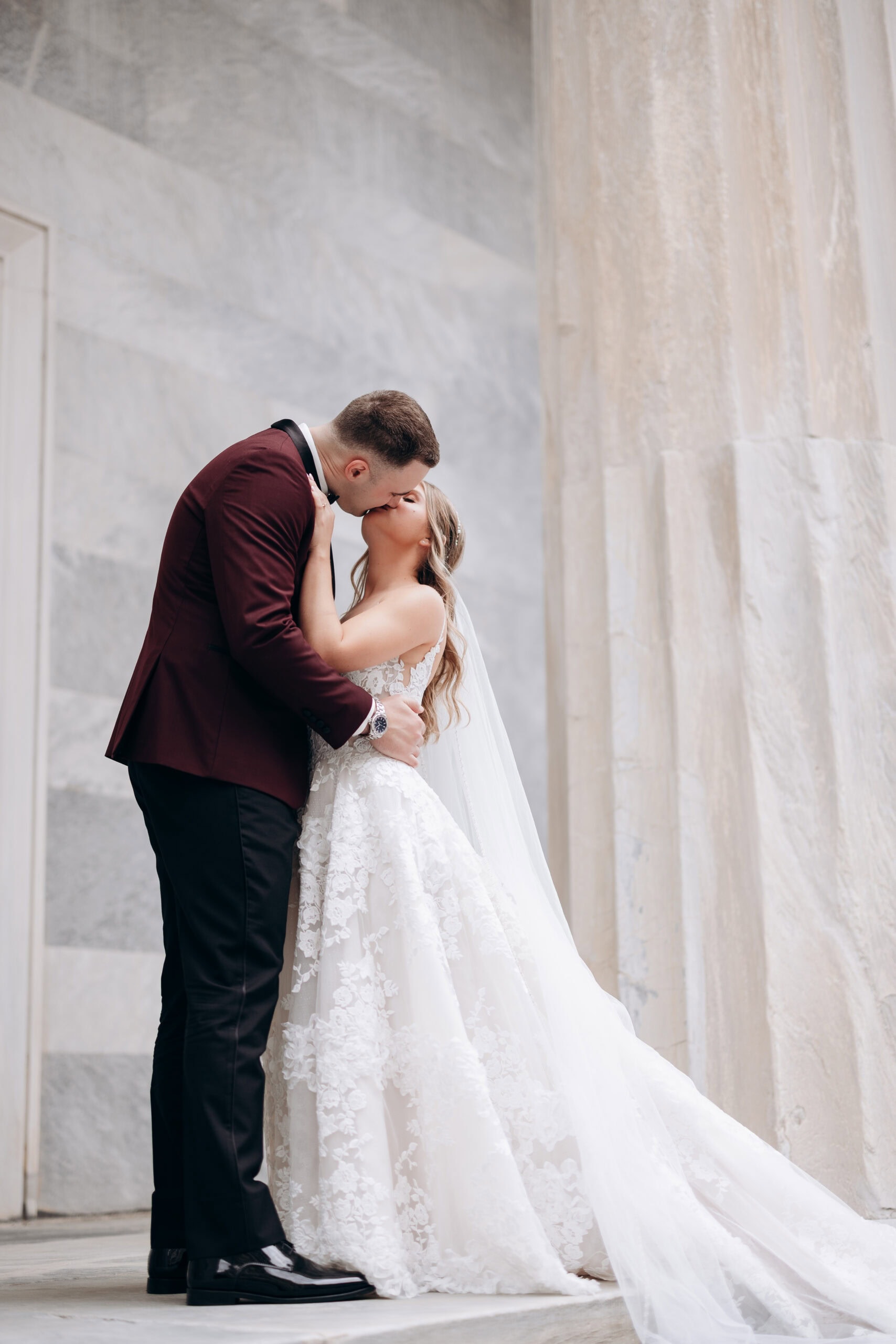 Bride and groom sharing a kiss beside marble columns at Union Trust, Philadelphia, PA, highlighting intricate lace gown and burgundy tuxedo.