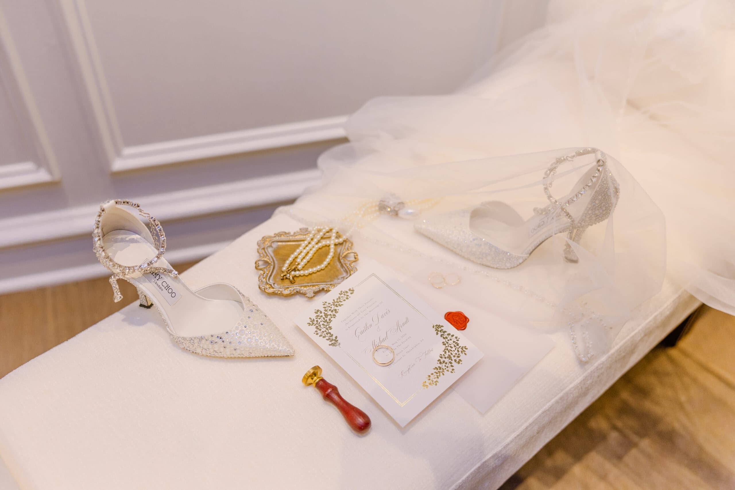 Crystal-embellished Jimmy Choo heels, pearl necklace on ornate tray, gold-foiled wedding invitation with wax seal, and wedding rings on white fabric surface.