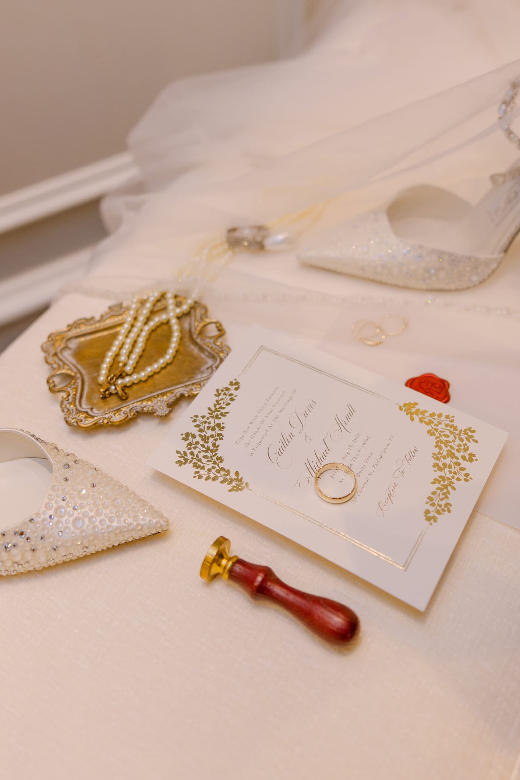 Close-up of pearl necklace on ornate tray, beaded bridal shoe, gold wedding rings on invitation, and wax seal stamp on cream fabric.
