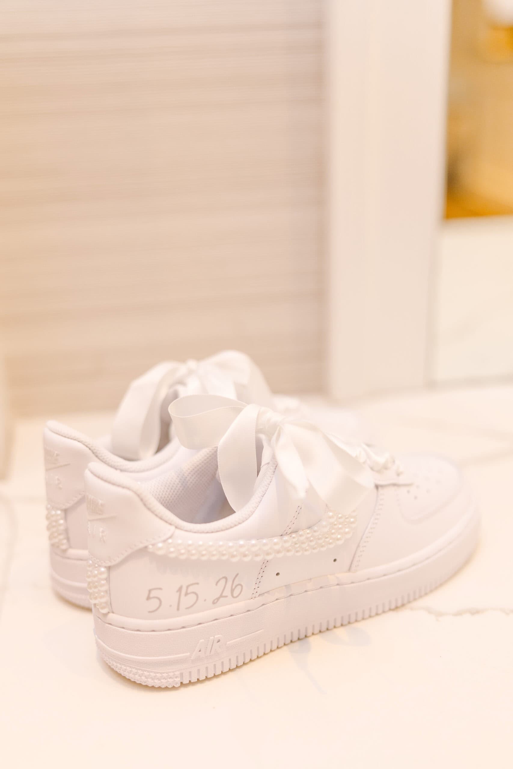 White Nike Air Force 1 sneakers decorated with pearls and satin ribbon laces, handwritten wedding date 5.15.26 on heel, on marble surface.