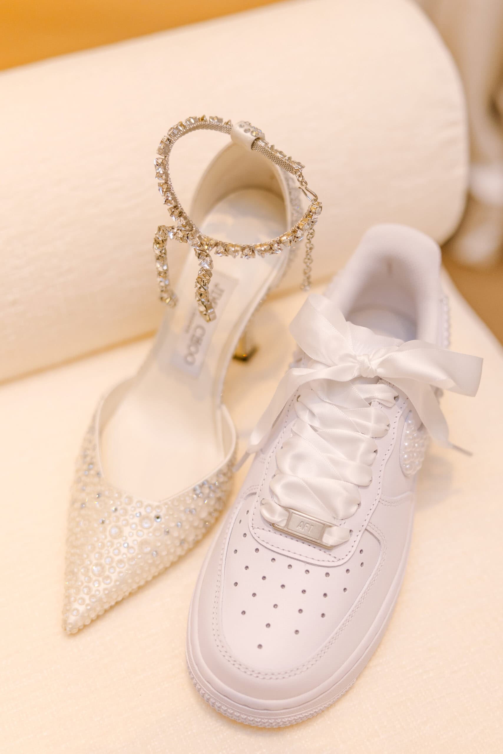 Close-up of a pearl and crystal-embellished Jimmy Choo high heel alongside a white Nike Air Force 1 sneaker with satin ribbon laces, placed on a textured cream surface.
