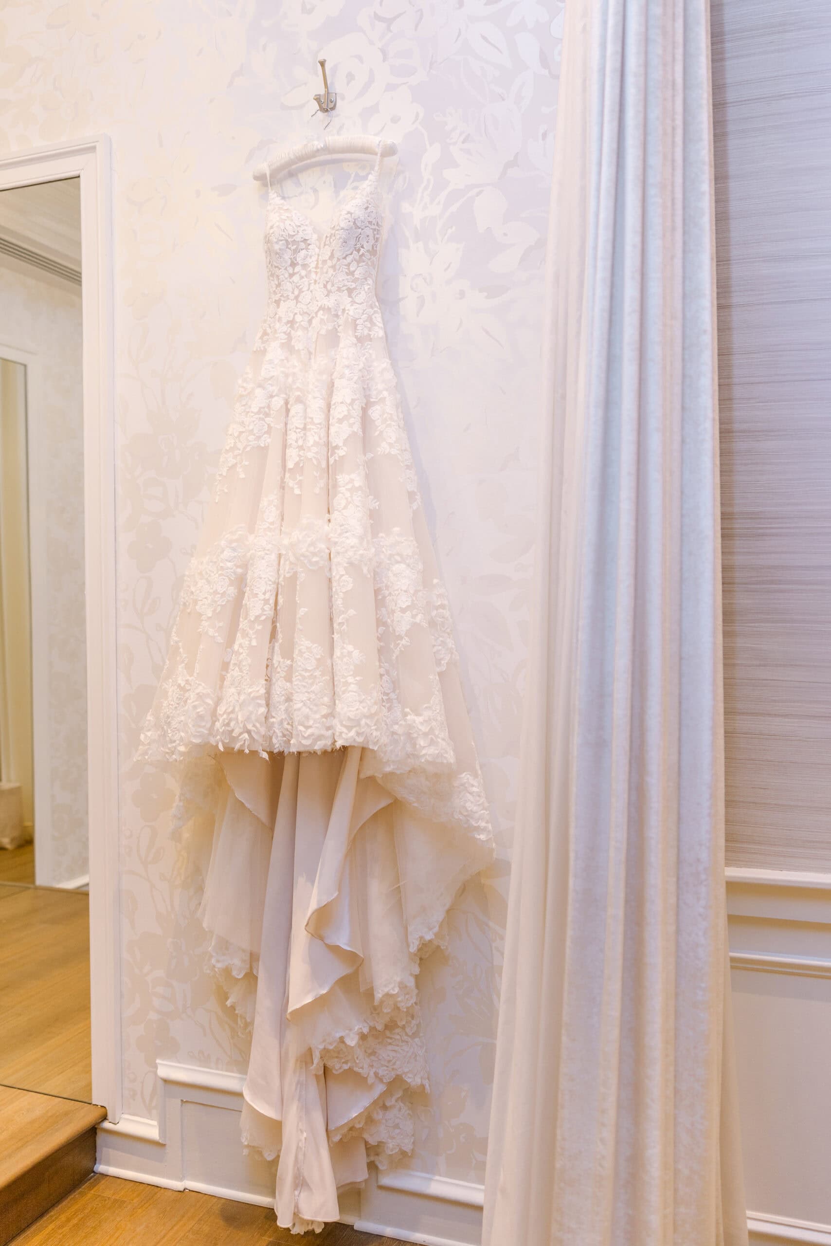 Detailed lace wedding dress hanging on a padded hanger against floral wallpaper near a mirror and curtain at Union Trust, Philadelphia.