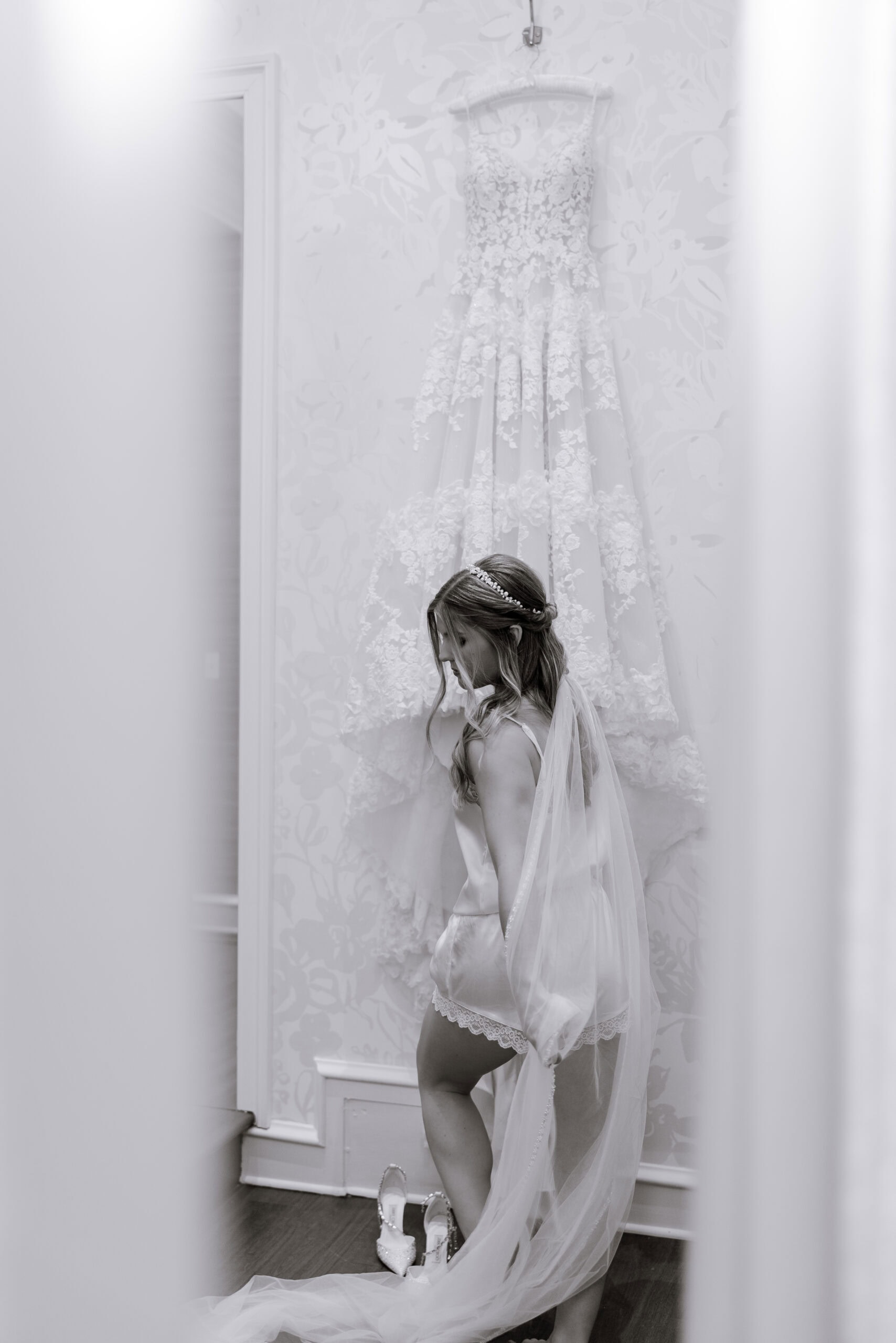 Bride in silk and lace lingerie with veil, standing beneath a hanging lace wedding gown in a softly lit room at Union Trust, Philadelphia.