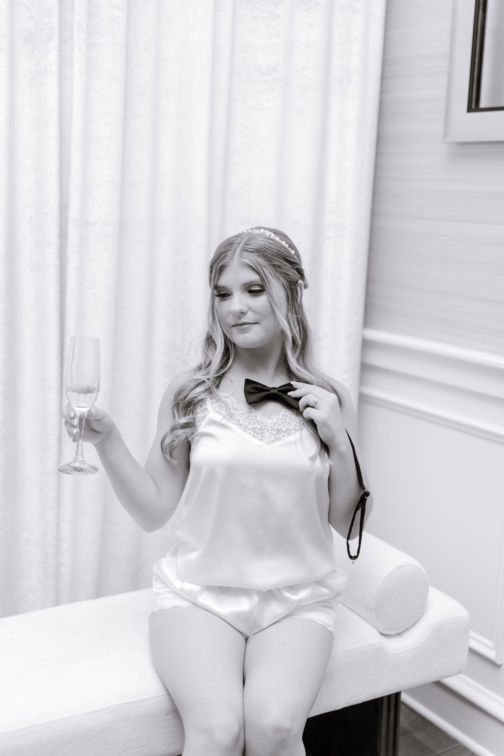 Bride seated on a white bench in silk lingerie holding a champagne flute and black bow tie in a softly lit room at Union Trust, Philadelphia.