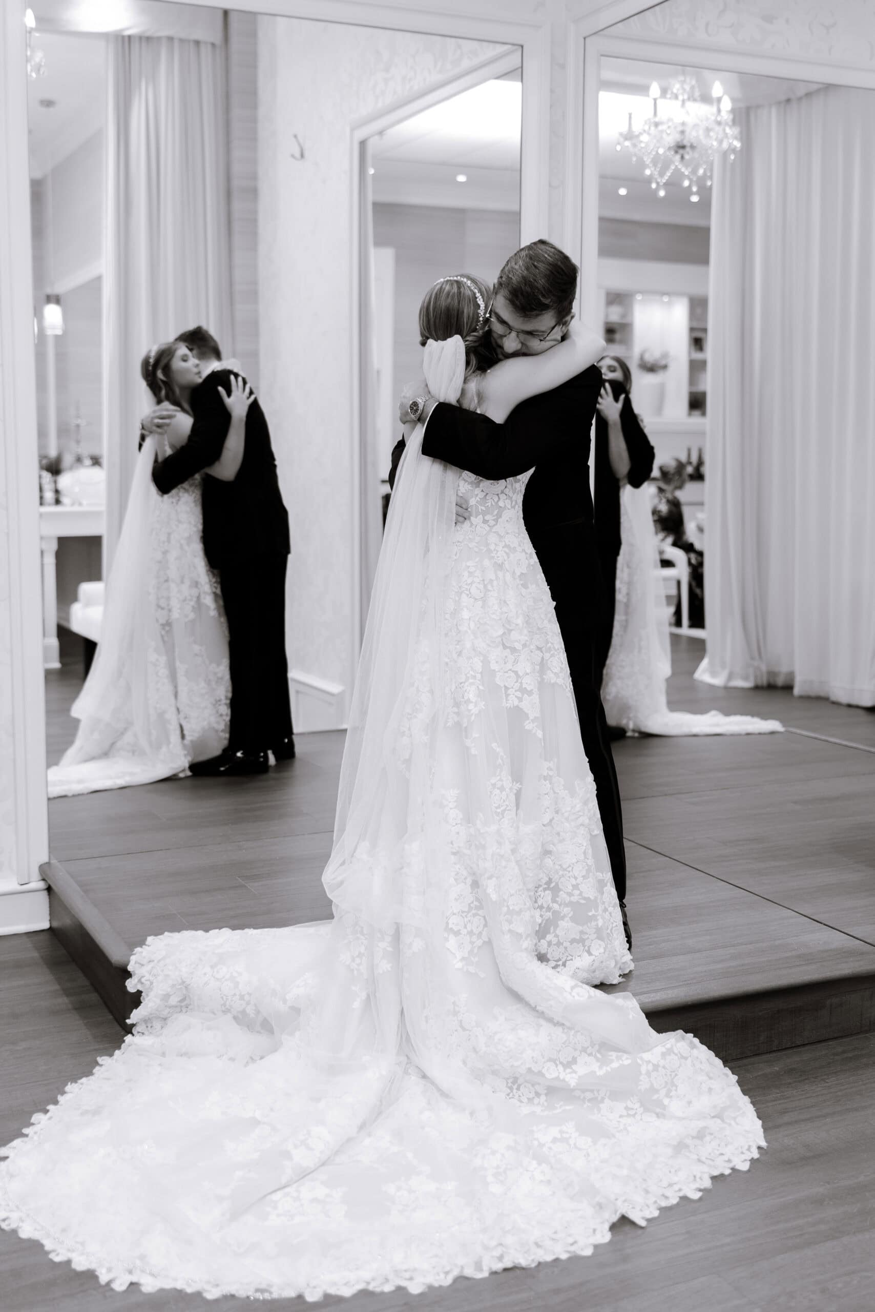 Bride and groom in an intimate embrace reflected in a large mirror, bride wearing a lace wedding gown with a long train and veil, groom in a dark suit, interior with chandelier and curtains at Union Trust, Philadelphia.