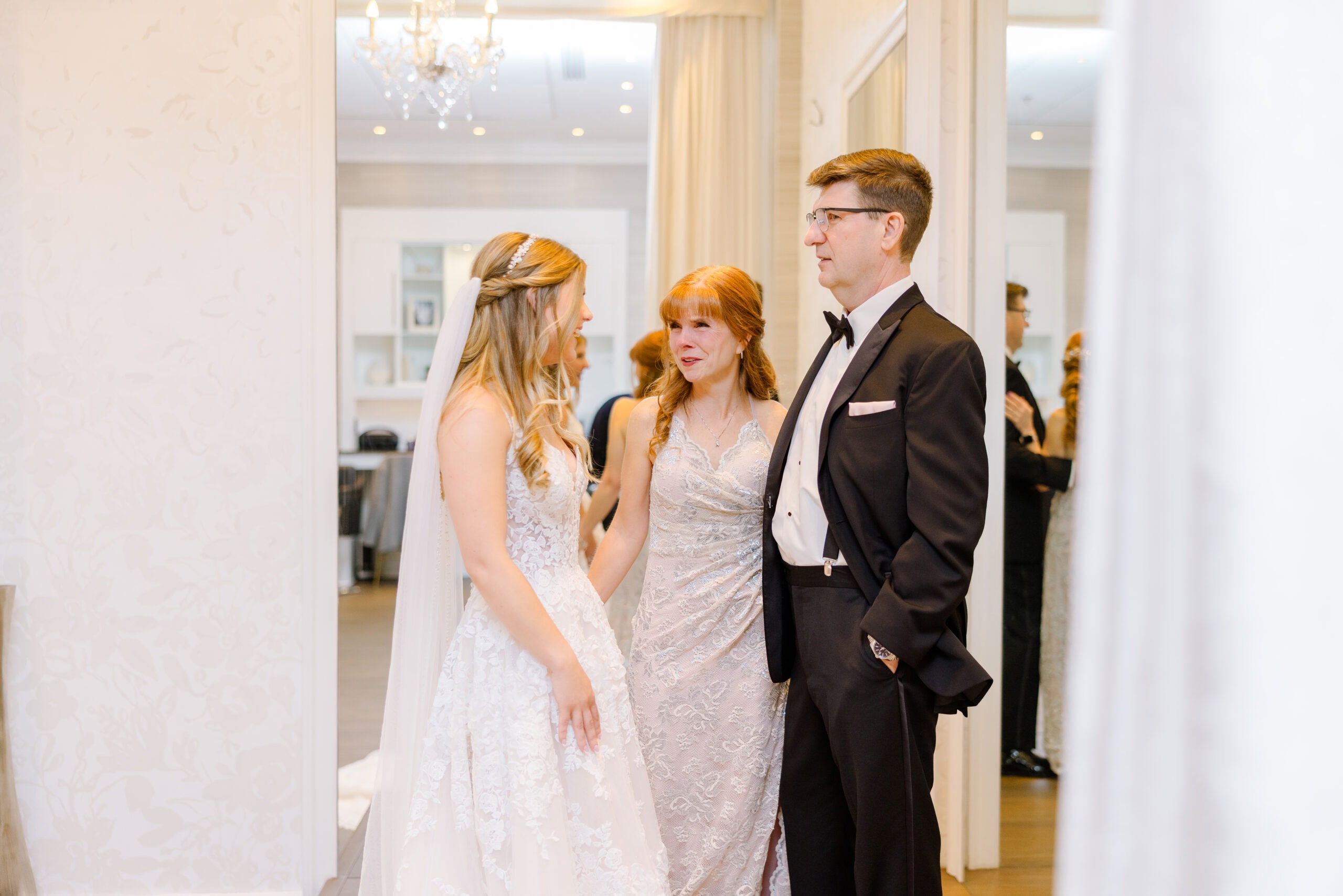 Bride in lace wedding dress with veil talking to her mother in a silver lace gown and father in black tuxedo inside a bright room at Union Trust Philadelphia.