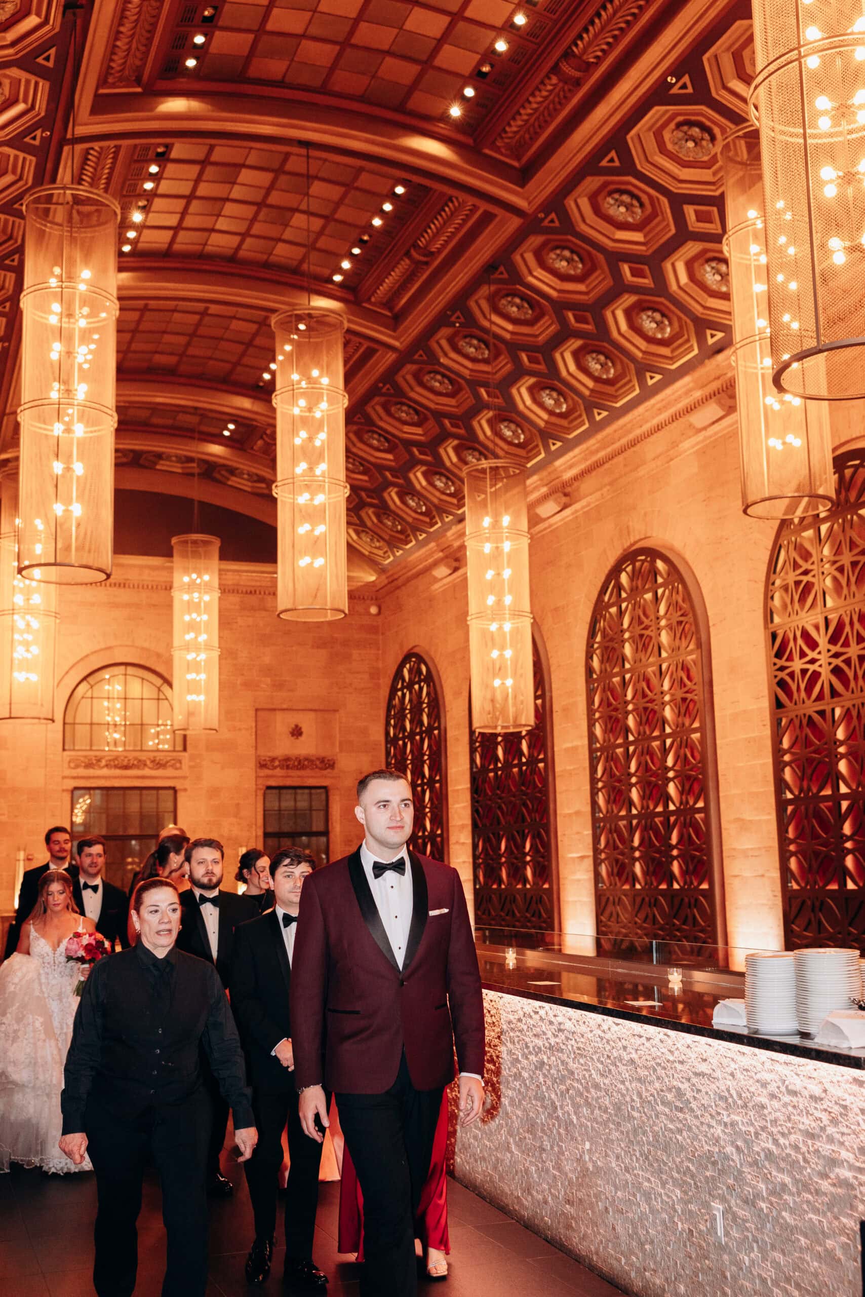Wedding procession led by groom in burgundy tuxedo walking through Union Trust Philadelphia with ornate ceiling and glowing cylindrical chandeliers