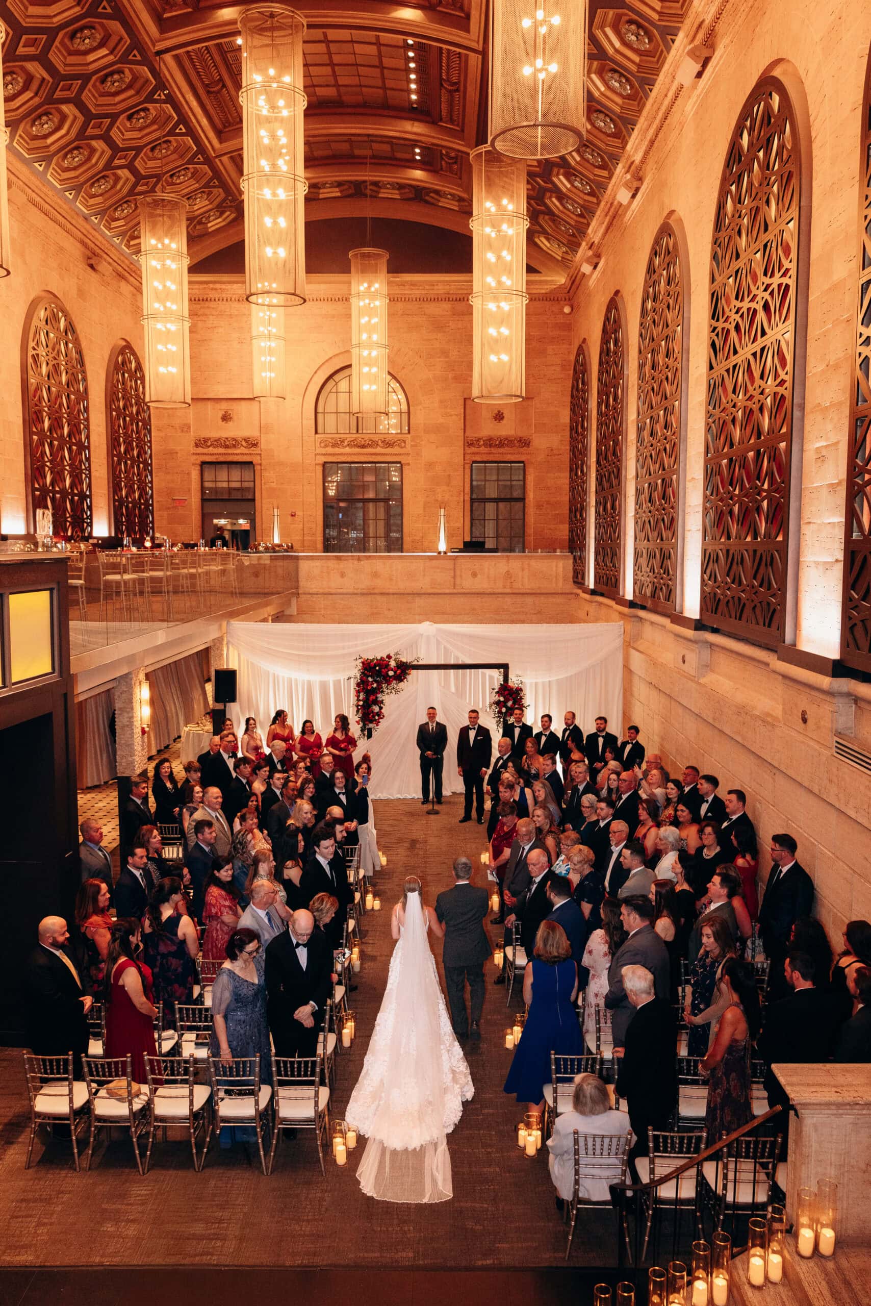 Bride walking down the aisle in an elegant hall with high ceilings, ornate windows, and hanging chandeliers at Union Trust, Philadelphia.