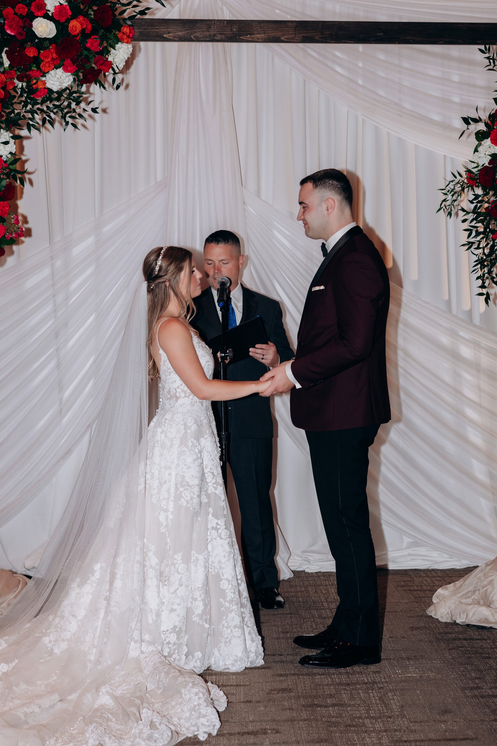 Bride and groom holding hands during wedding ceremony with officiant, floral arrangements, and white draped backdrop at Union Trust Philadelphia.
