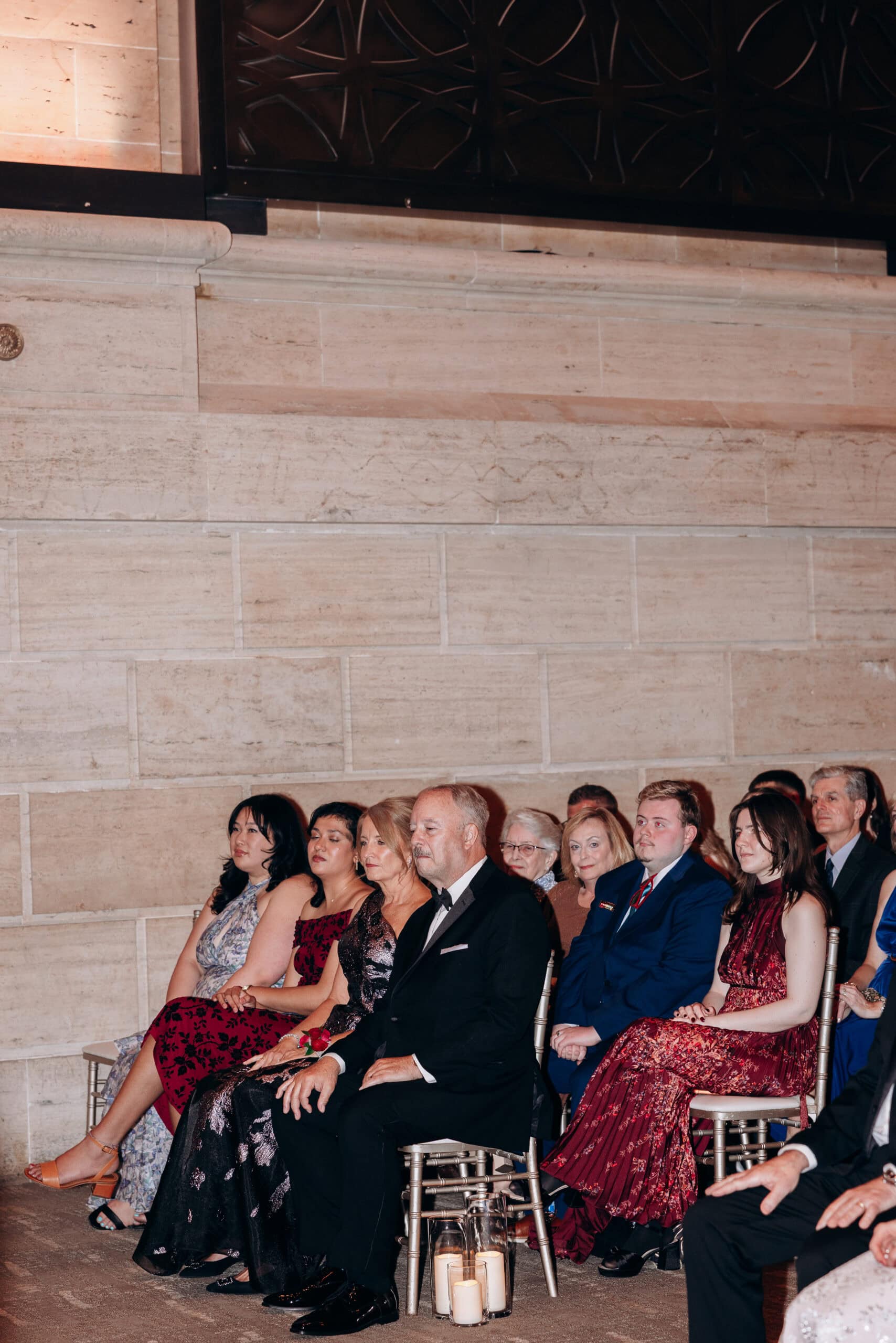 Guests in formal wear seated attentively on chiavari chairs with candles on the floor, against a stone wall at Union Trust, Philadelphia.