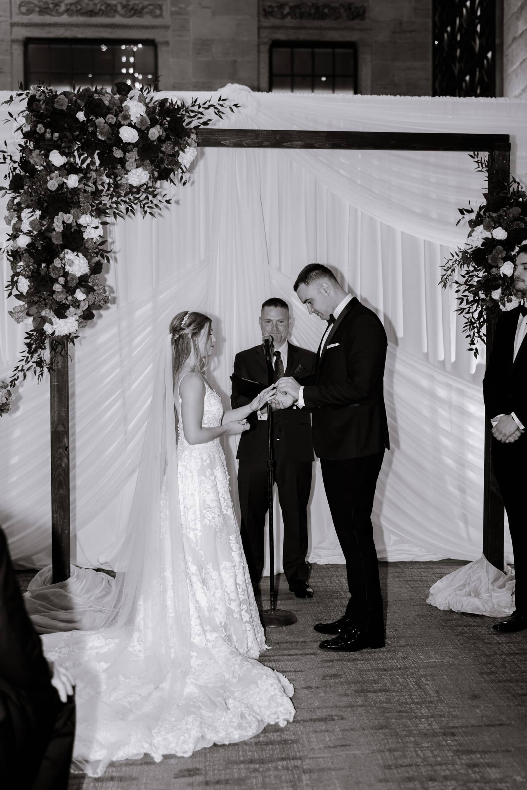 Bride and groom exchanging rings during wedding ceremony under floral wooden arch with officiant at Union Trust, Philadelphia.
