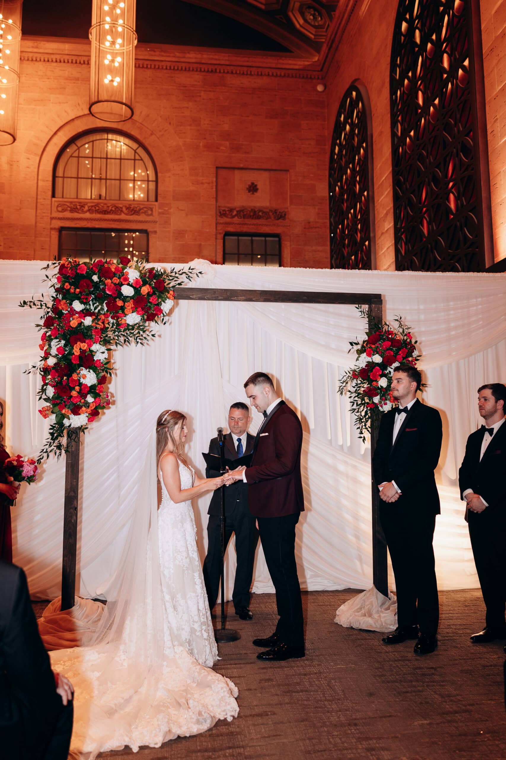 Bride and groom exchanging rings under a wooden arch draped with white fabric and red floral arrangements inside Union Trust, Philadelphia.