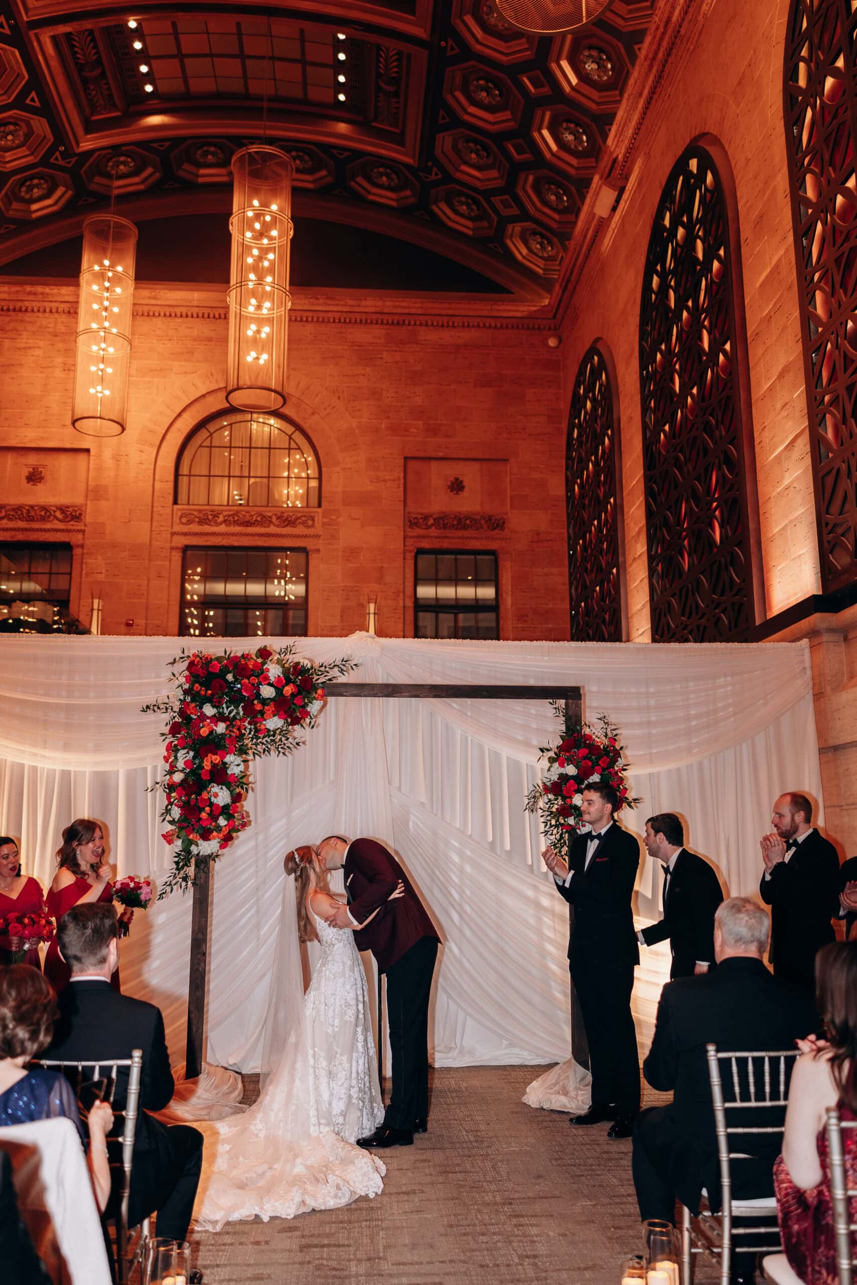 Bride and groom sharing a kiss under a floral arch during their wedding ceremony inside Union Trust, Philadelphia, with guests applauding.