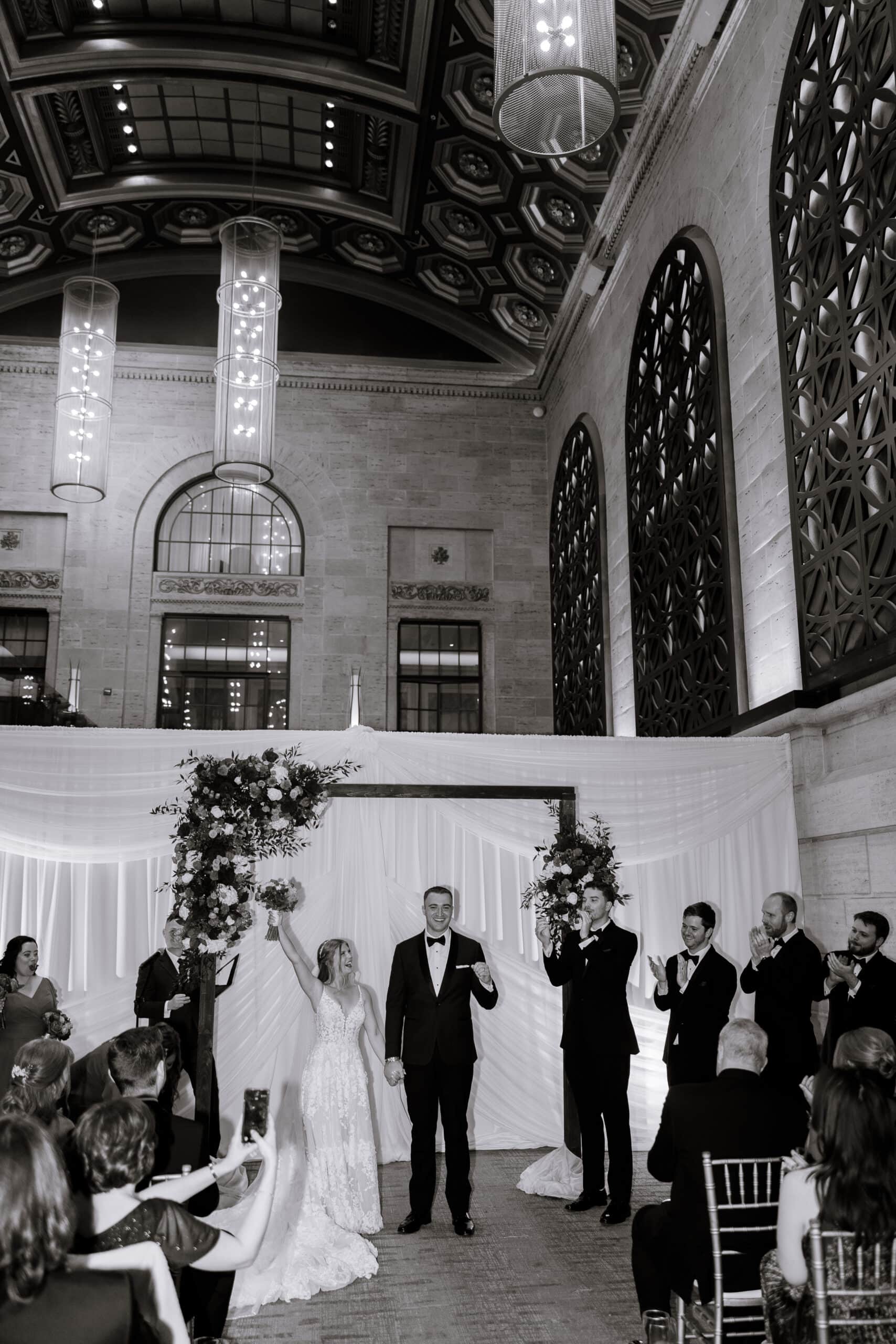 Bride and groom holding hands and celebrating at their wedding ceremony under floral arch and intricate ceiling at Union Trust, Philadelphia.