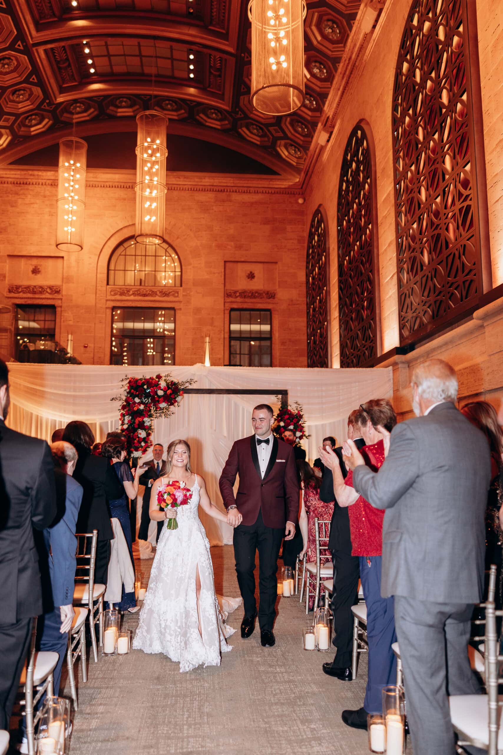 Bride and groom walking down the aisle at Union Trust Philadelphia with guests applauding, ornate ceiling and warm lighting overhead.