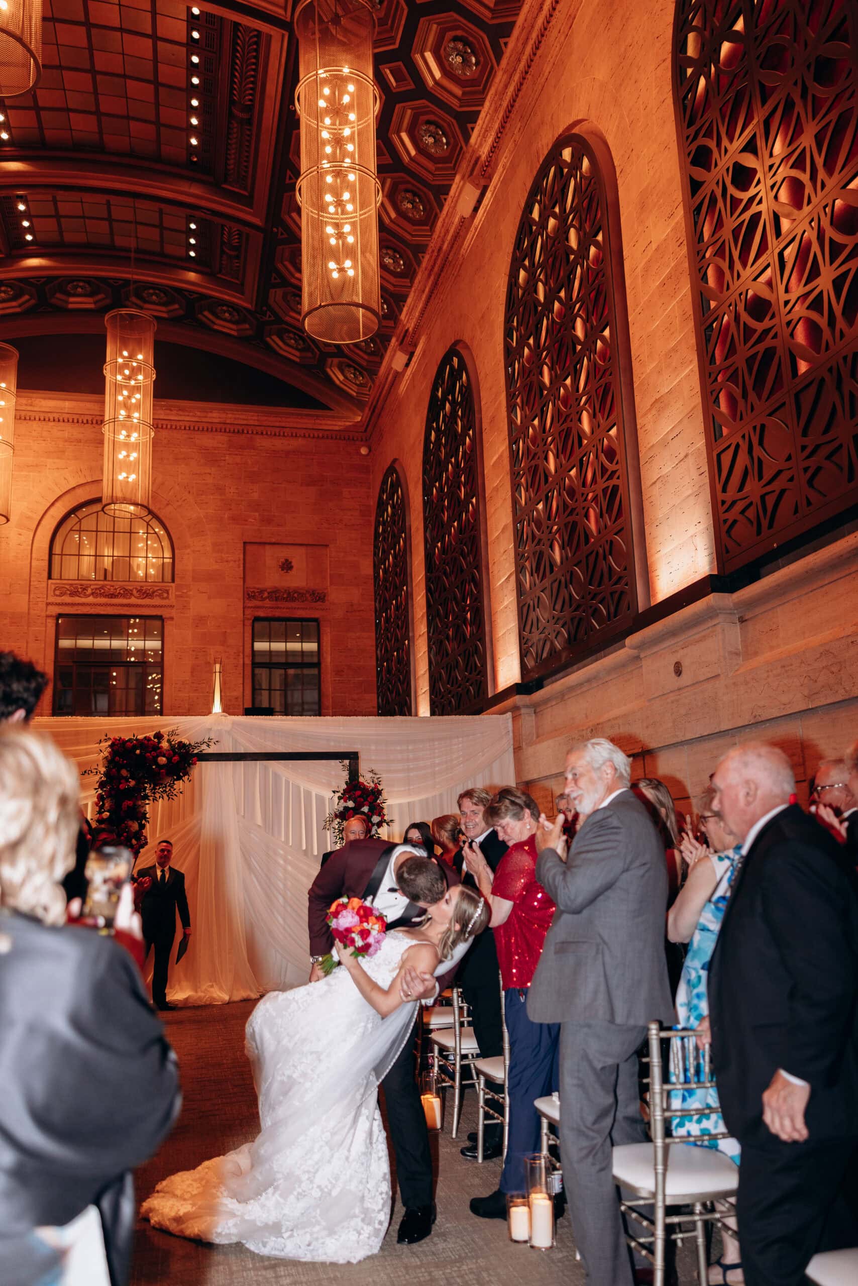 Groom dipping bride for a kiss during wedding ceremony at Union Trust, Philadelphia, with guests applauding and ornate ceiling overhead.